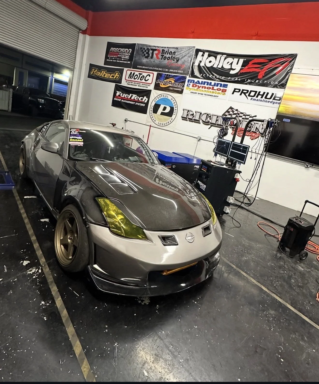 A silver race car with black and yellow details parked inside a garage with banners and equipment on the walls.