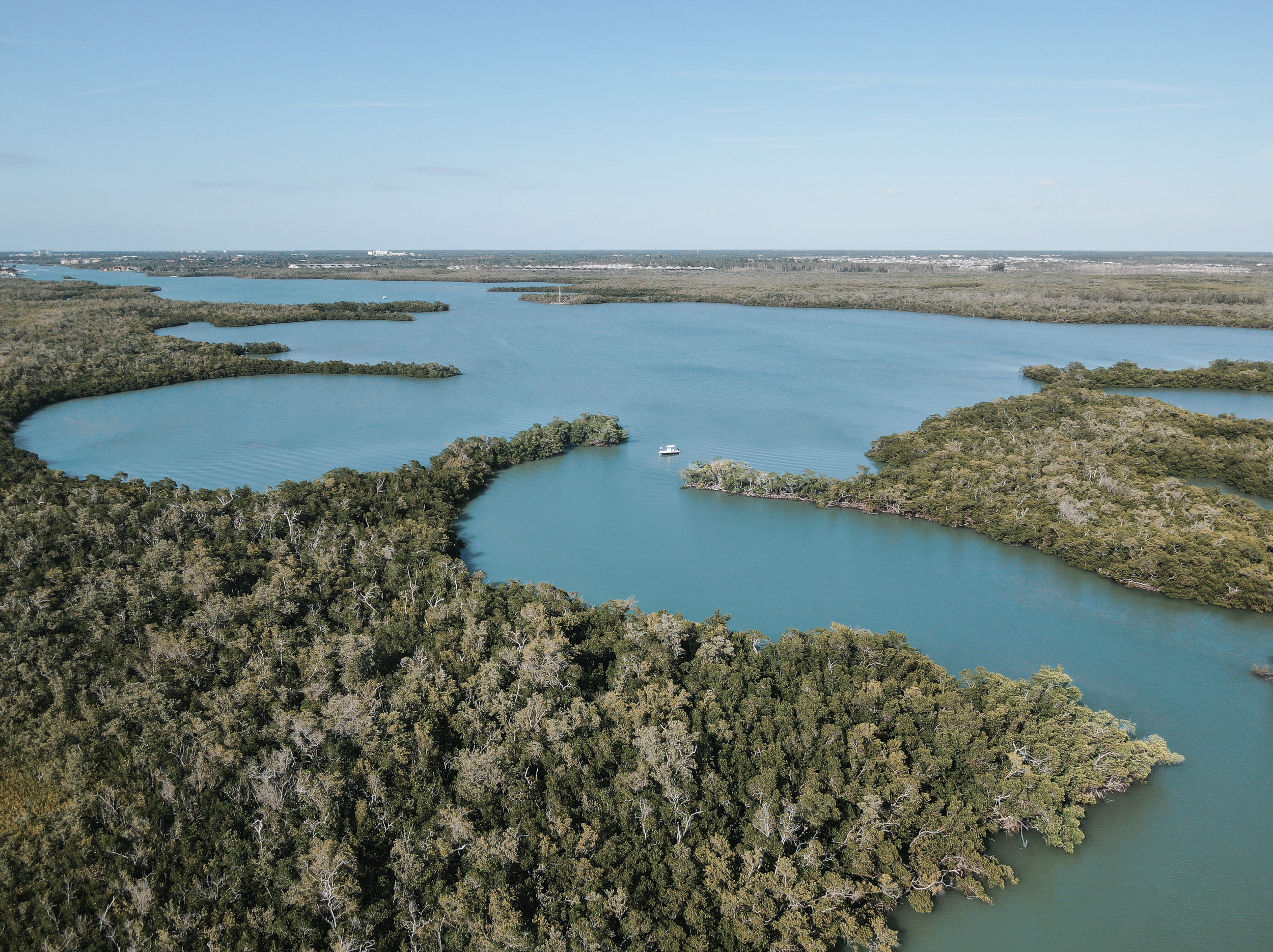 Aerial view of a large lake surrounded by dense green forest, with a small boat on the water and a distant horizon under a clear blue sky.