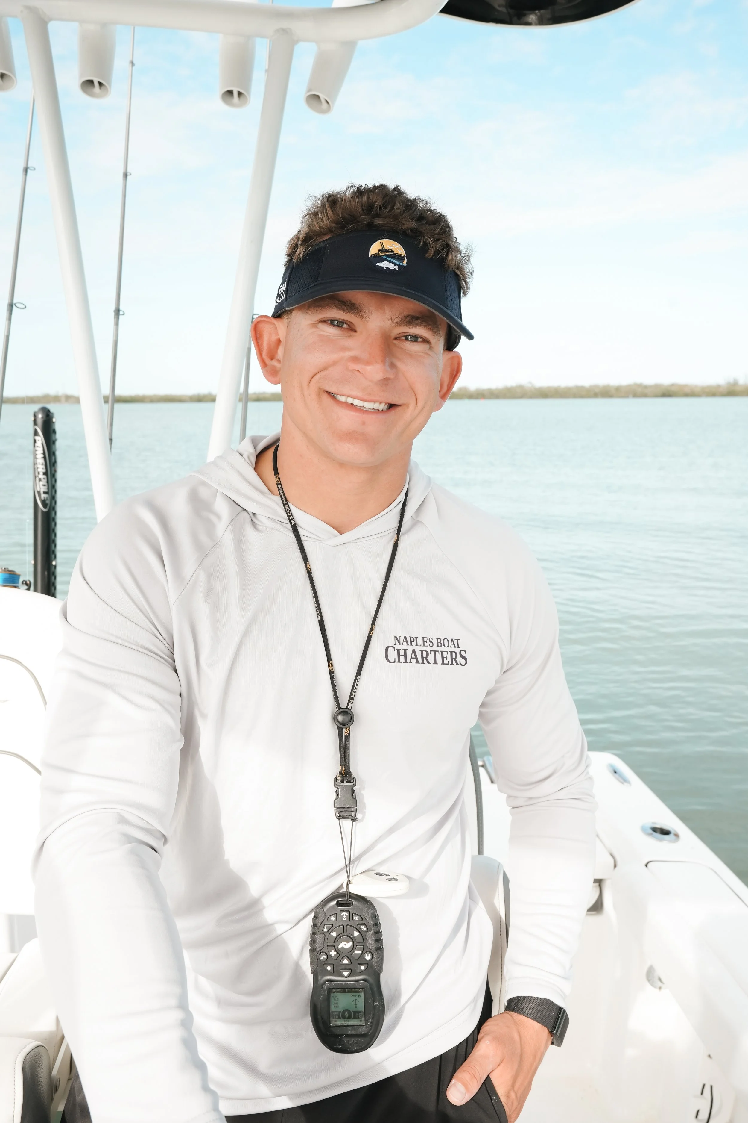 A smiling man wearing a white long-sleeve shirt with 'Naples Boat Charters' logo, a black visor, and a watch, standing on a boat with water and a cloudy sky in the background.