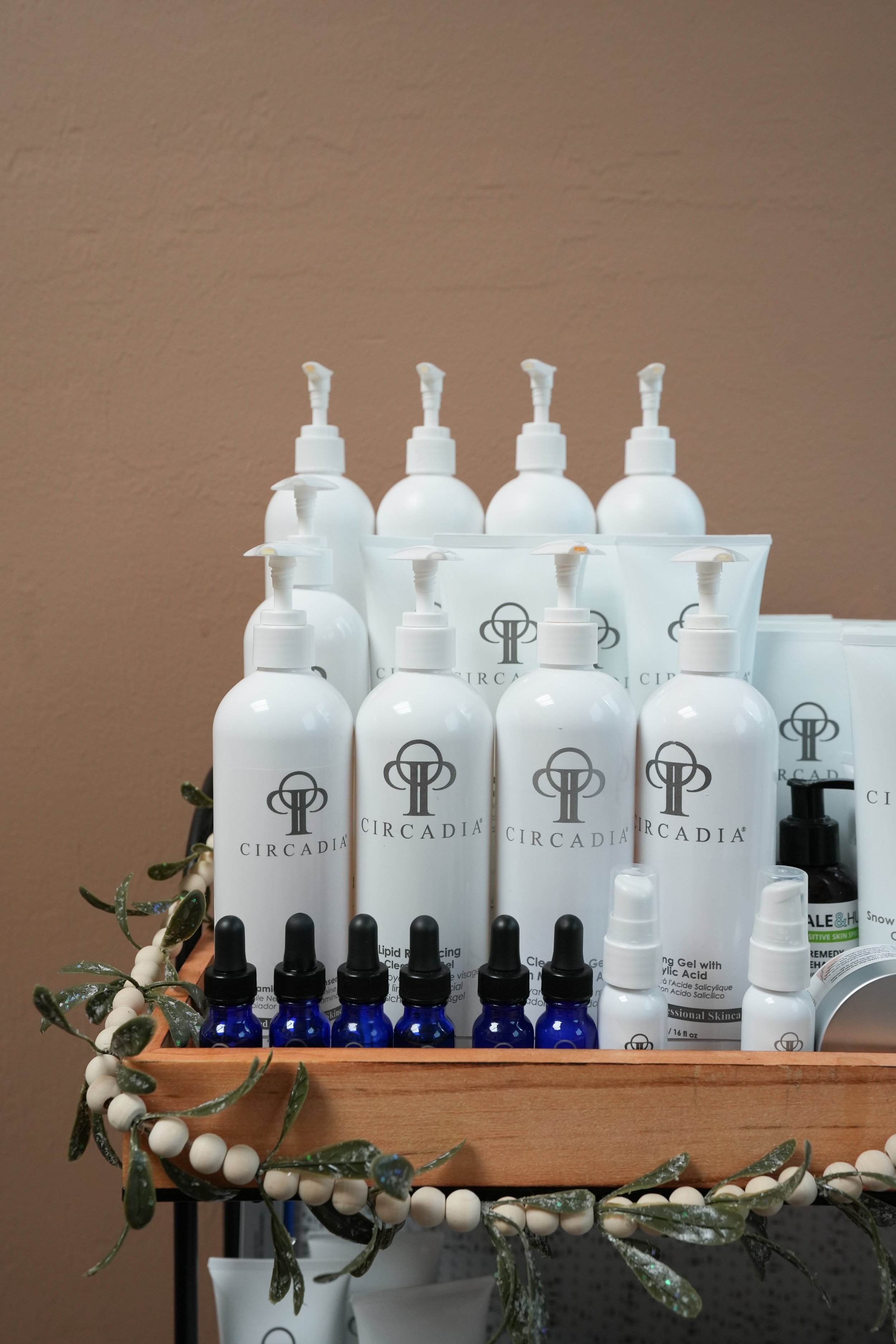 Display of skincare products, including white bottles with pump dispensers, small blue bottles with droppers, and other containers, arranged on a wooden shelf decorated with white and green bead garland, against a beige wall background.