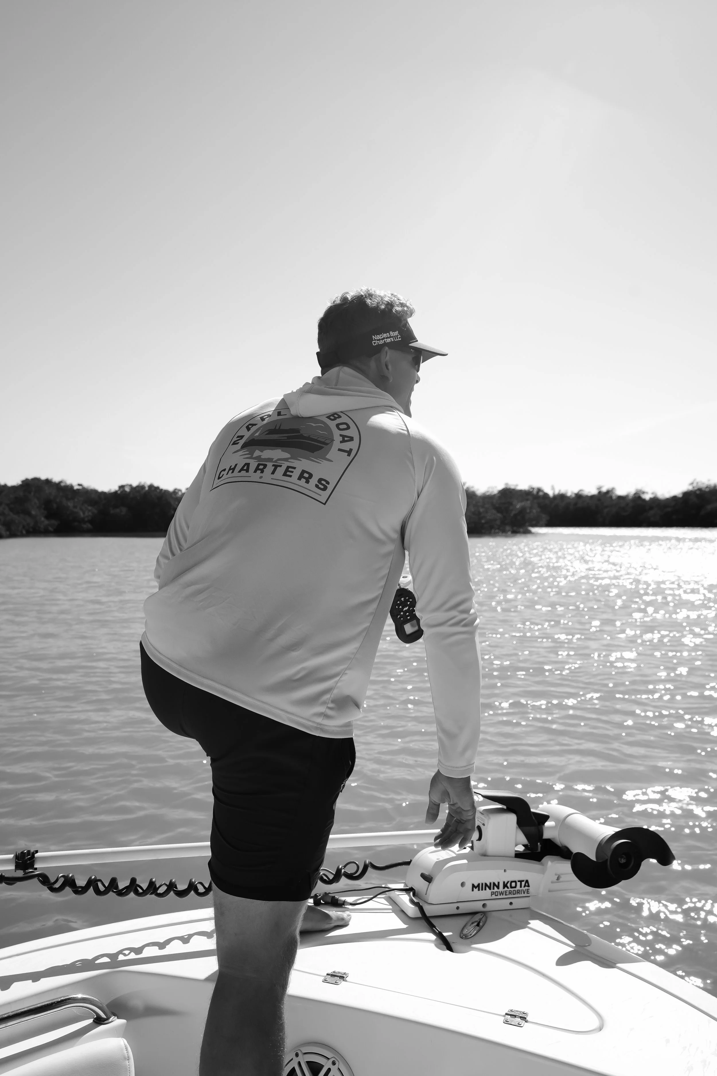 Man standing on a boat near the water, holding a Minn Kota Power Drive trolling motor, wearing a cap and jacket with 'Naples Boat Charters' logo, looking out towards the water.