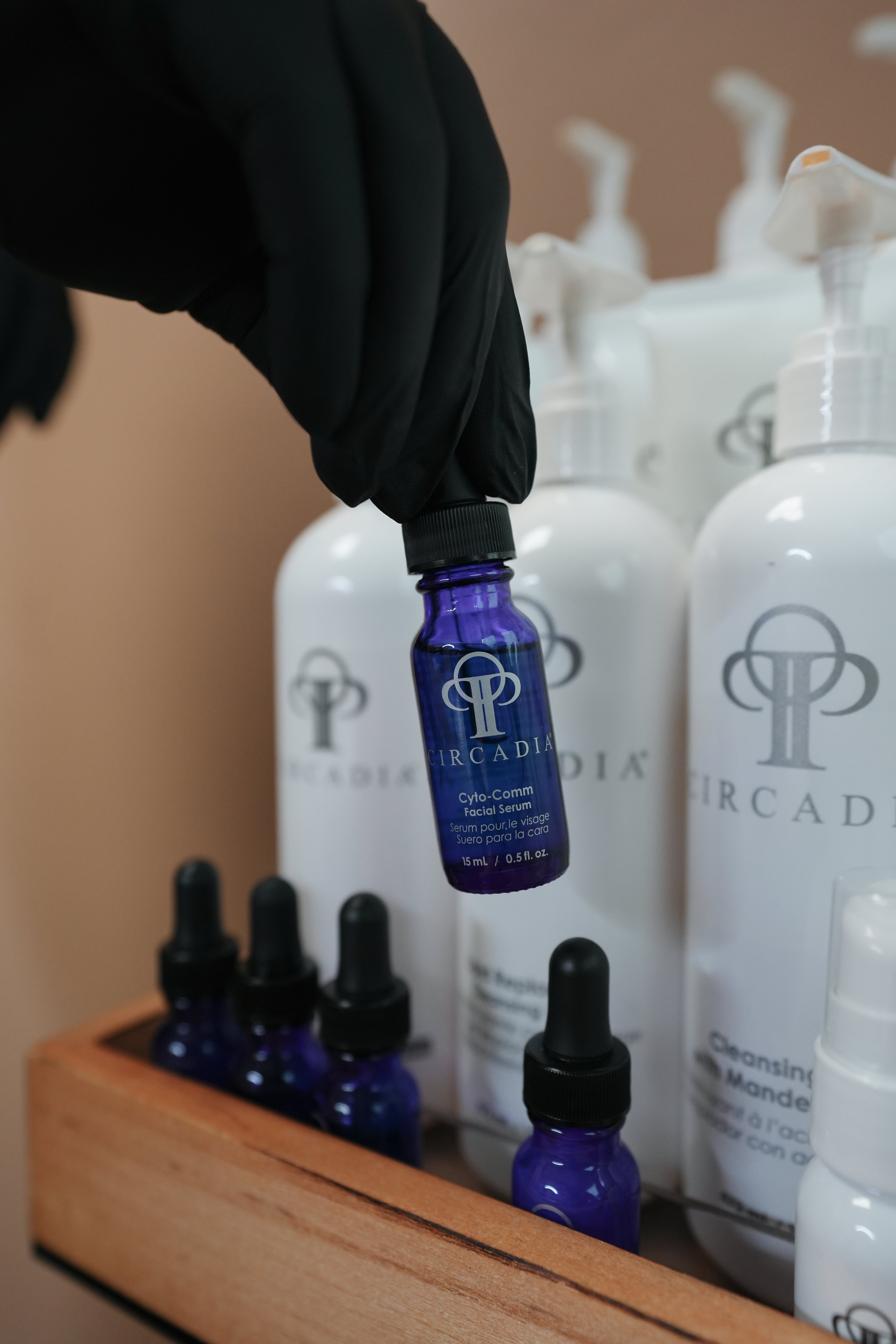 Person wearing black gloves holding a small blue serum bottle labeled CircaDia Cyto-Comm Facial Serum, with larger white bottles and smaller blue serum bottles on a wooden shelf in the background.