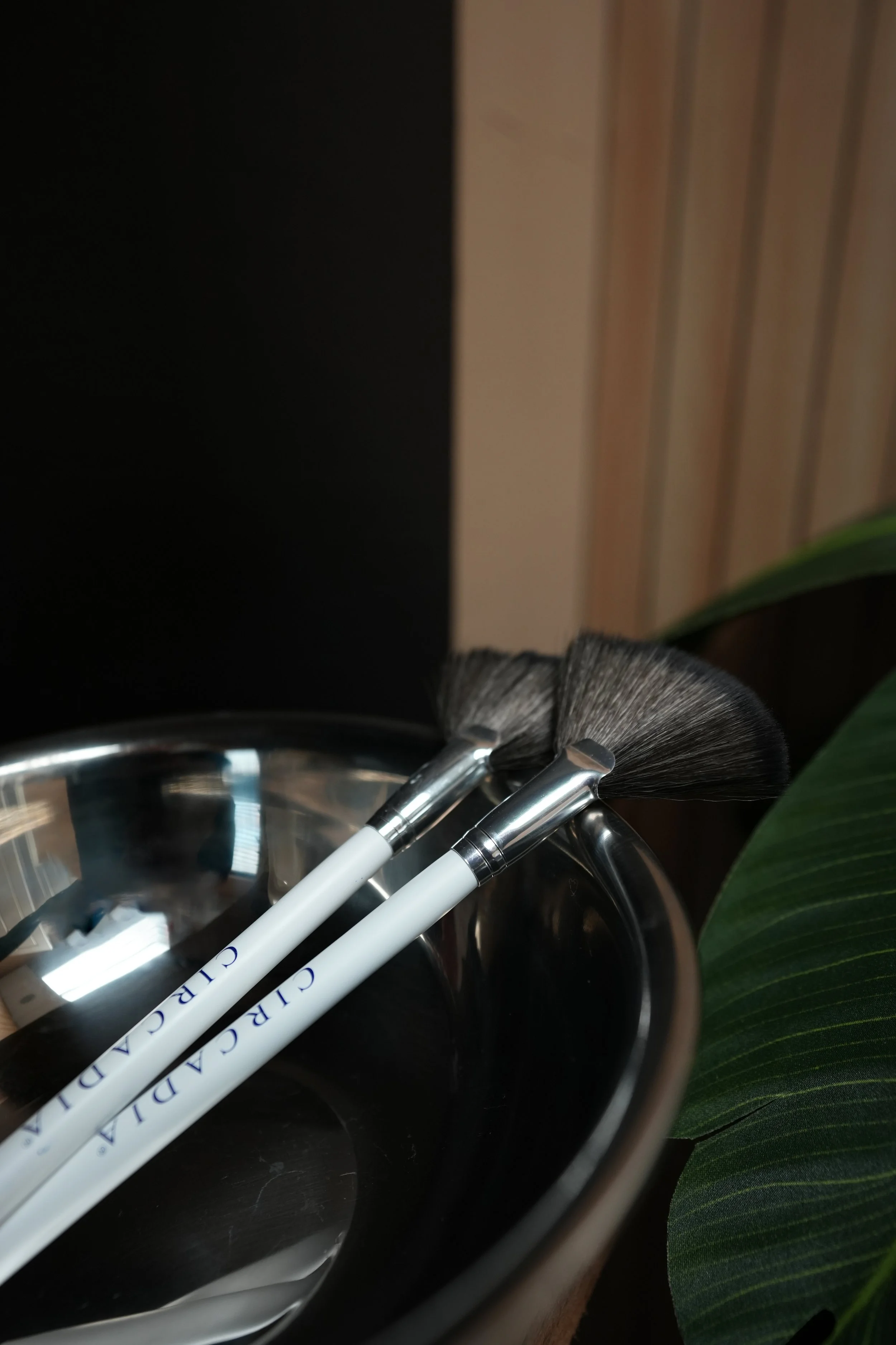 Two makeup brushes resting in a round copper container, with a green leafy plant partially visible in the foreground.