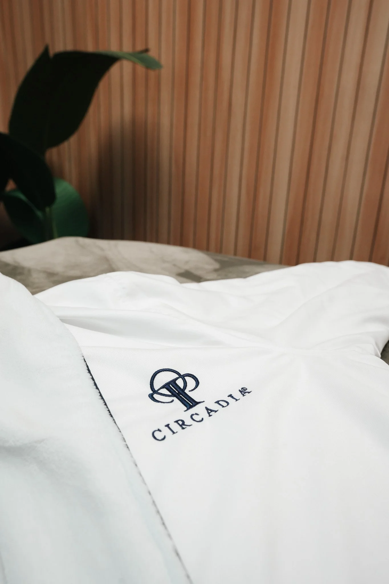 Close-up of a white embroidered linen with the Circa D'A logo, laid on a bed with a wooden panel wall and a green plant in the background.