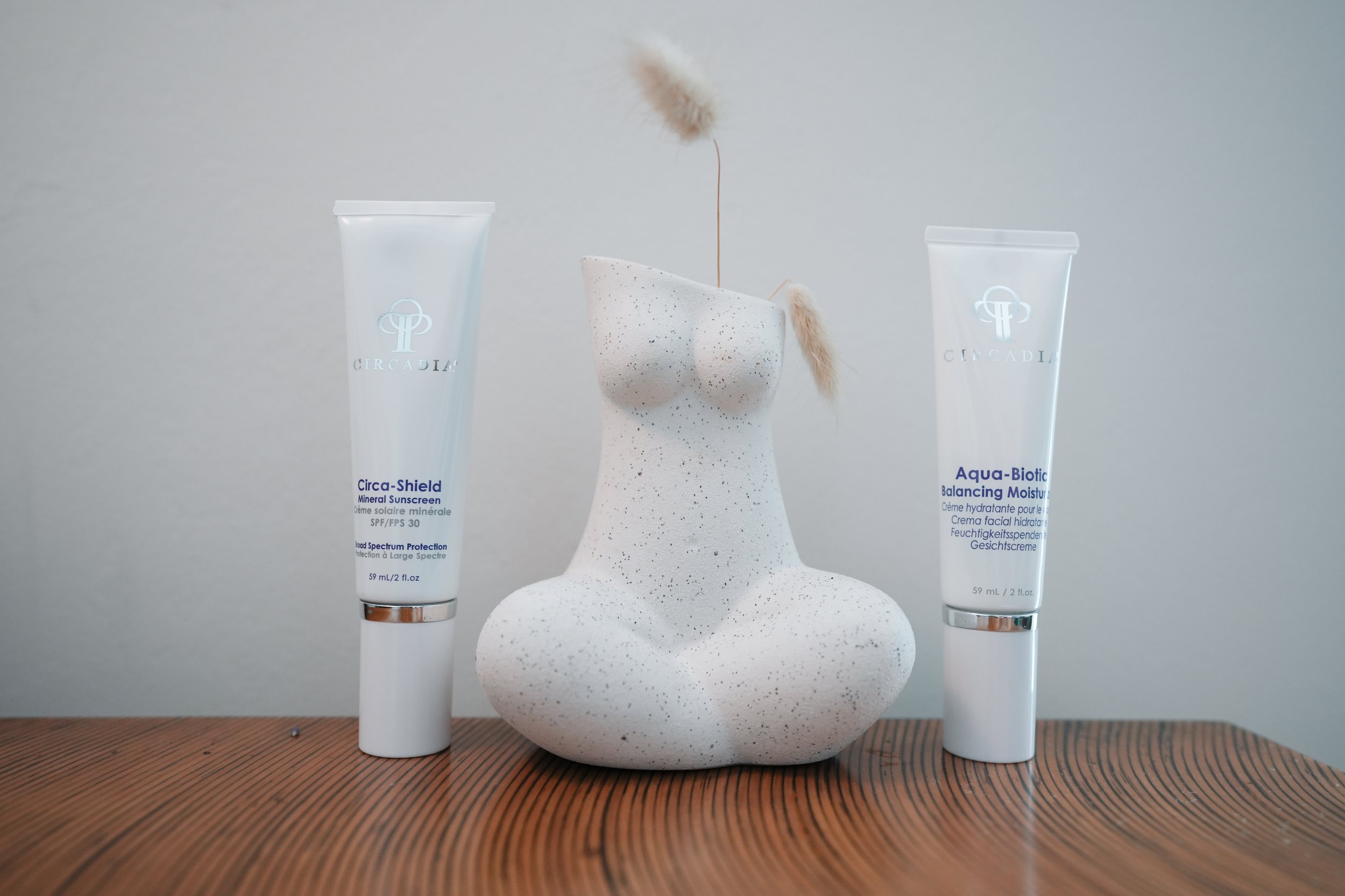 Decorative white vase in the shape of a female torso with dried pampas grass, flanked by two skincare tubes, on a wooden surface.