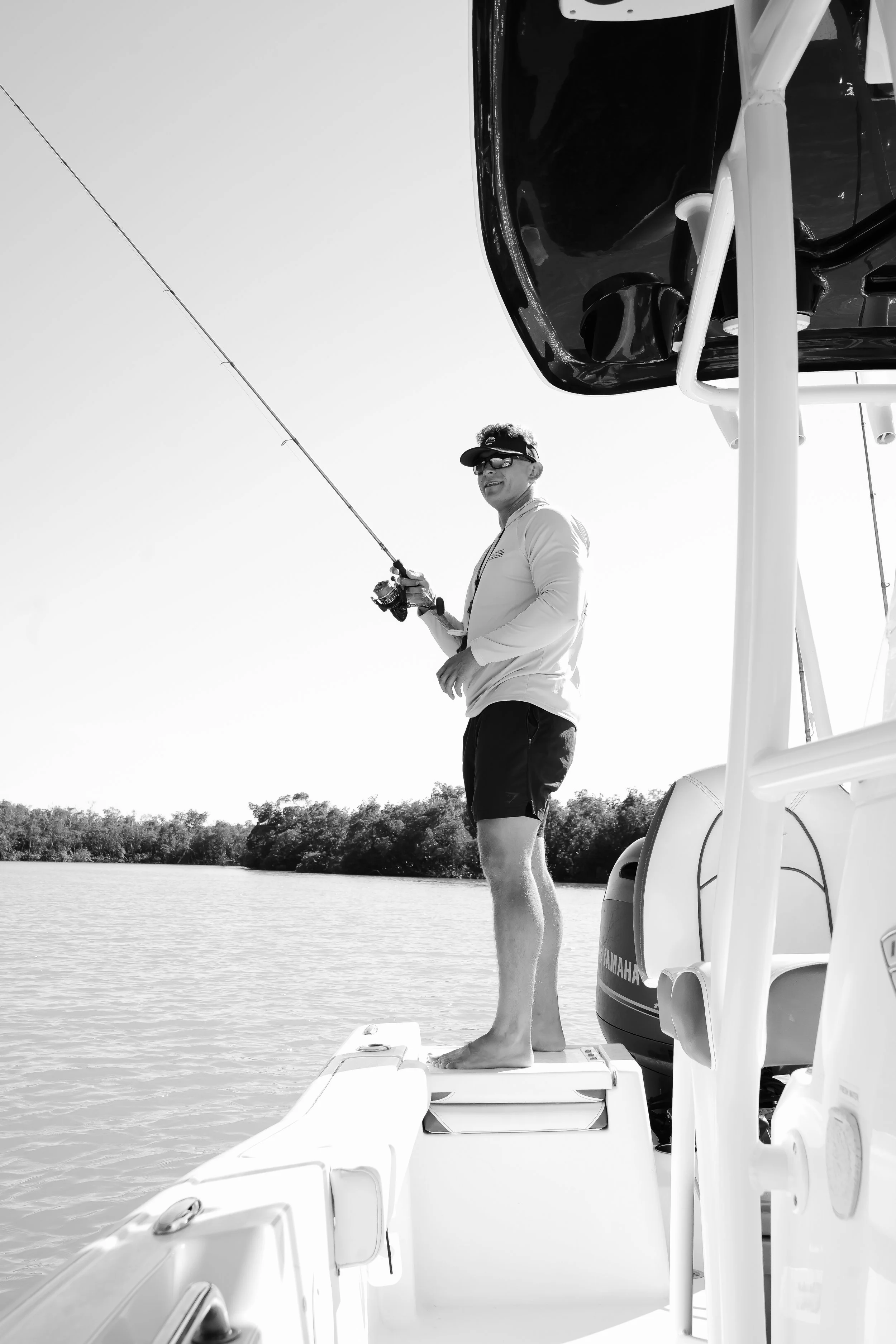 A man standing on the edge of a boat, fishing in a body of water on a sunny day, with trees in the background, wearing sunglasses, a long-sleeve shirt, and shorts.