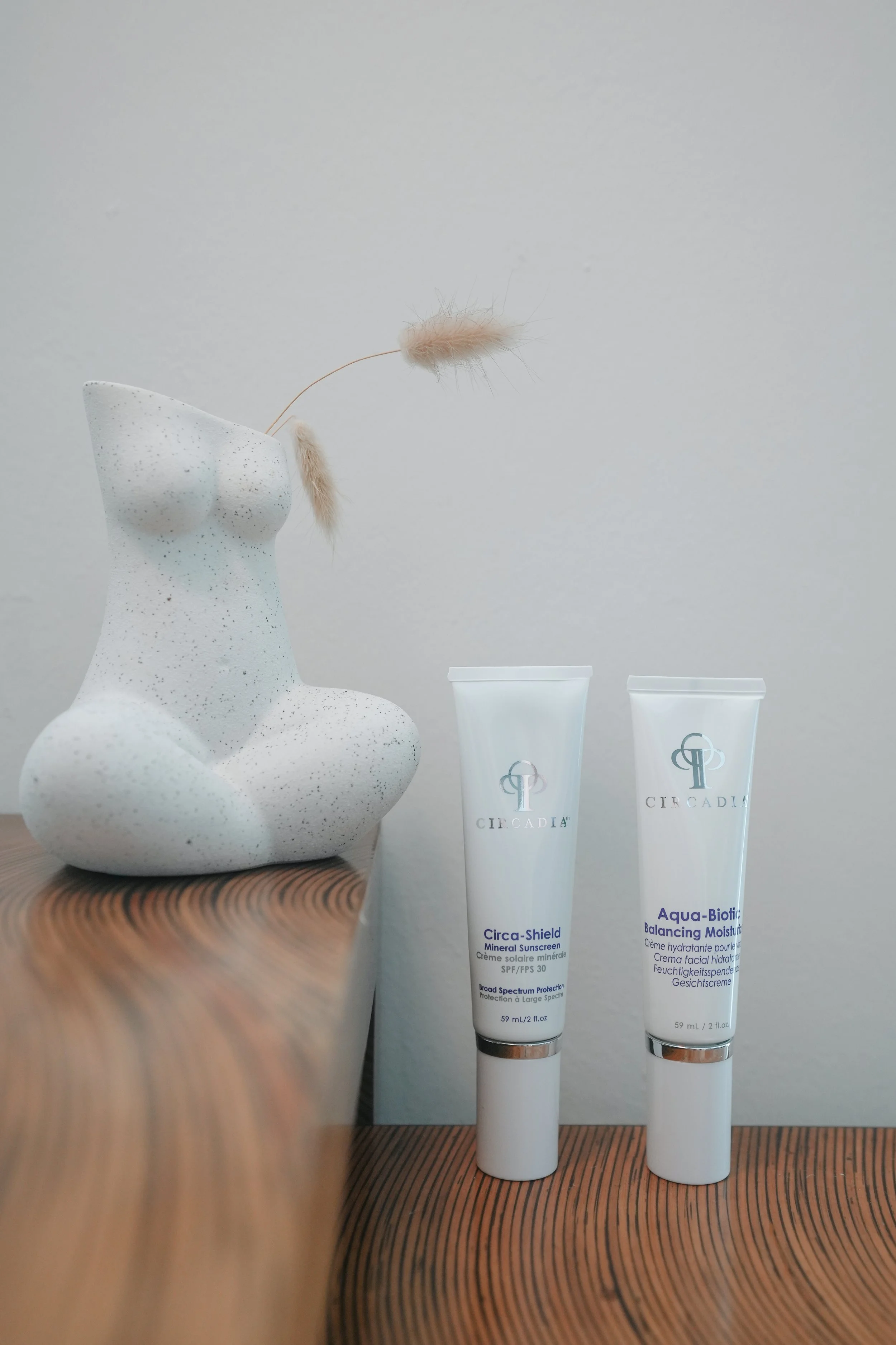 Decorative sculpture of a woman's torso with a fuzzy plant and two skincare tubes labeled Circa. One tube is labeled Circa-Shield and the other Aqua-Biotic Balancing Moisturizer, on a wooden surface against a plain wall.