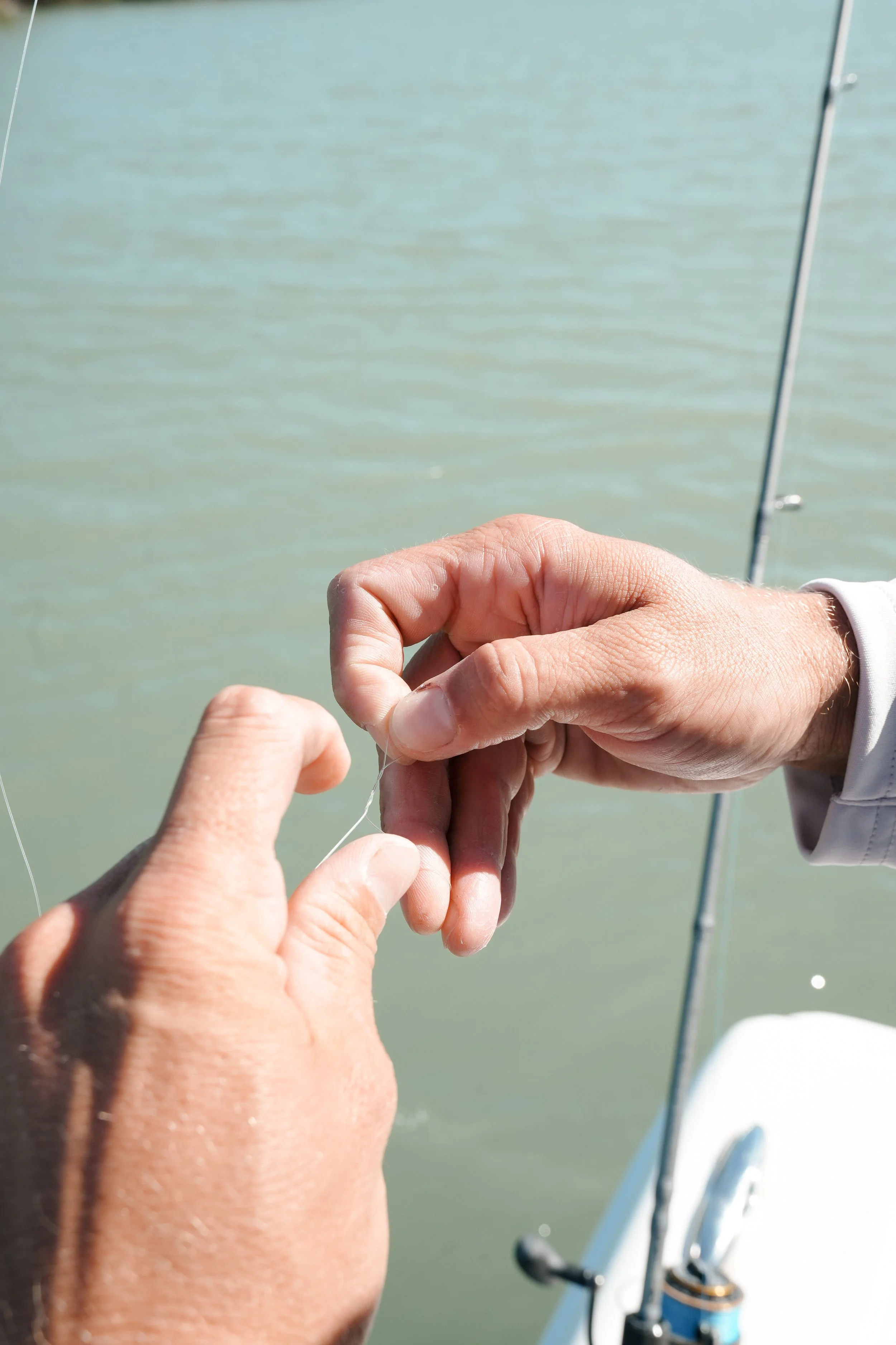 Close-up of two hands tying a fishing knot on a fishing line over a body of water, with a fishing rod in the background.