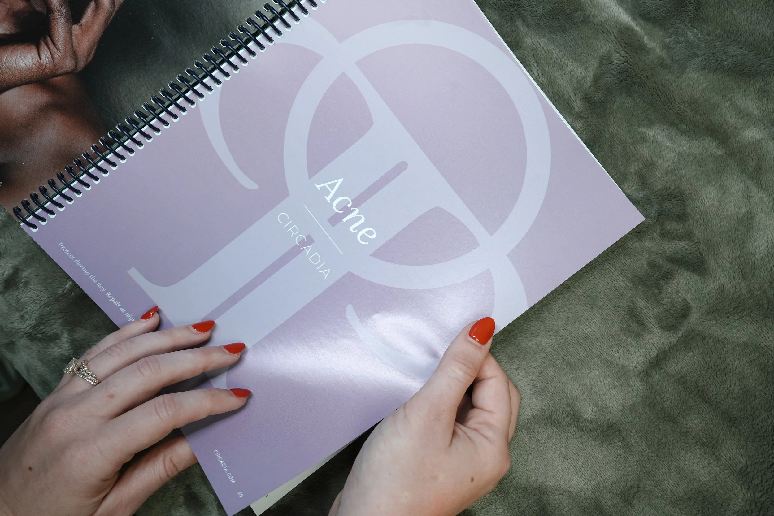 A person with red nail polish and a ring on their ring finger is holding a purple spiral-bound booklet with the words "Ache Circadia" on the cover, resting on a green textured surface.