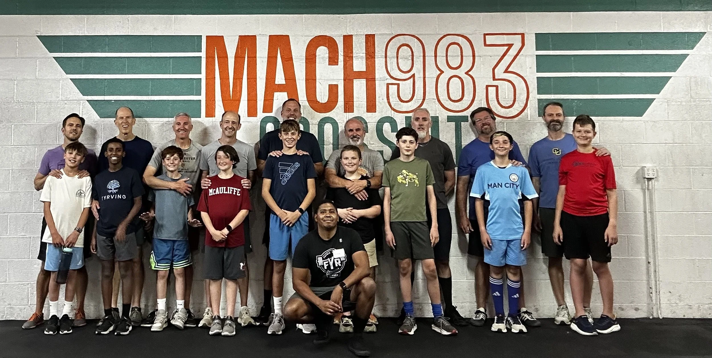 Group of children and adults posing together in front of a wall with painted text reading "MACH 98 3 POST". The group includes boys in athletic clothing, some wearing soccer jerseys, and adults standing behind them, all smiling.