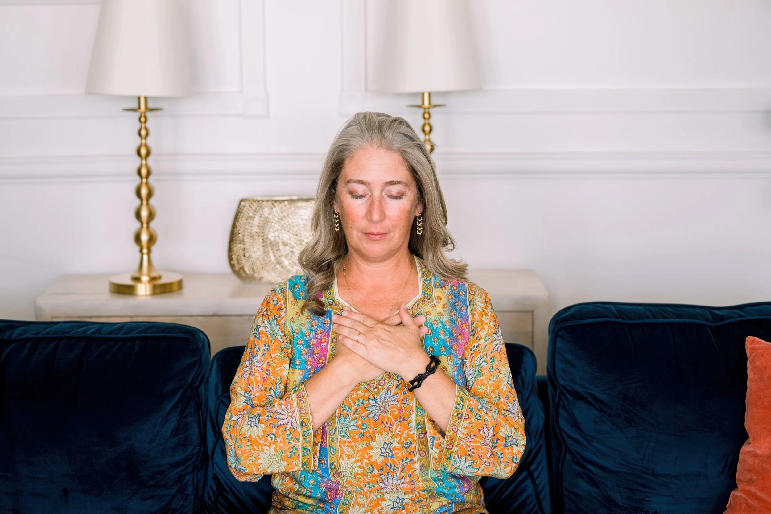 Woman sitting on a blue sofa with eyes closed and hands over her heart, illustrating Reiki self-practice.