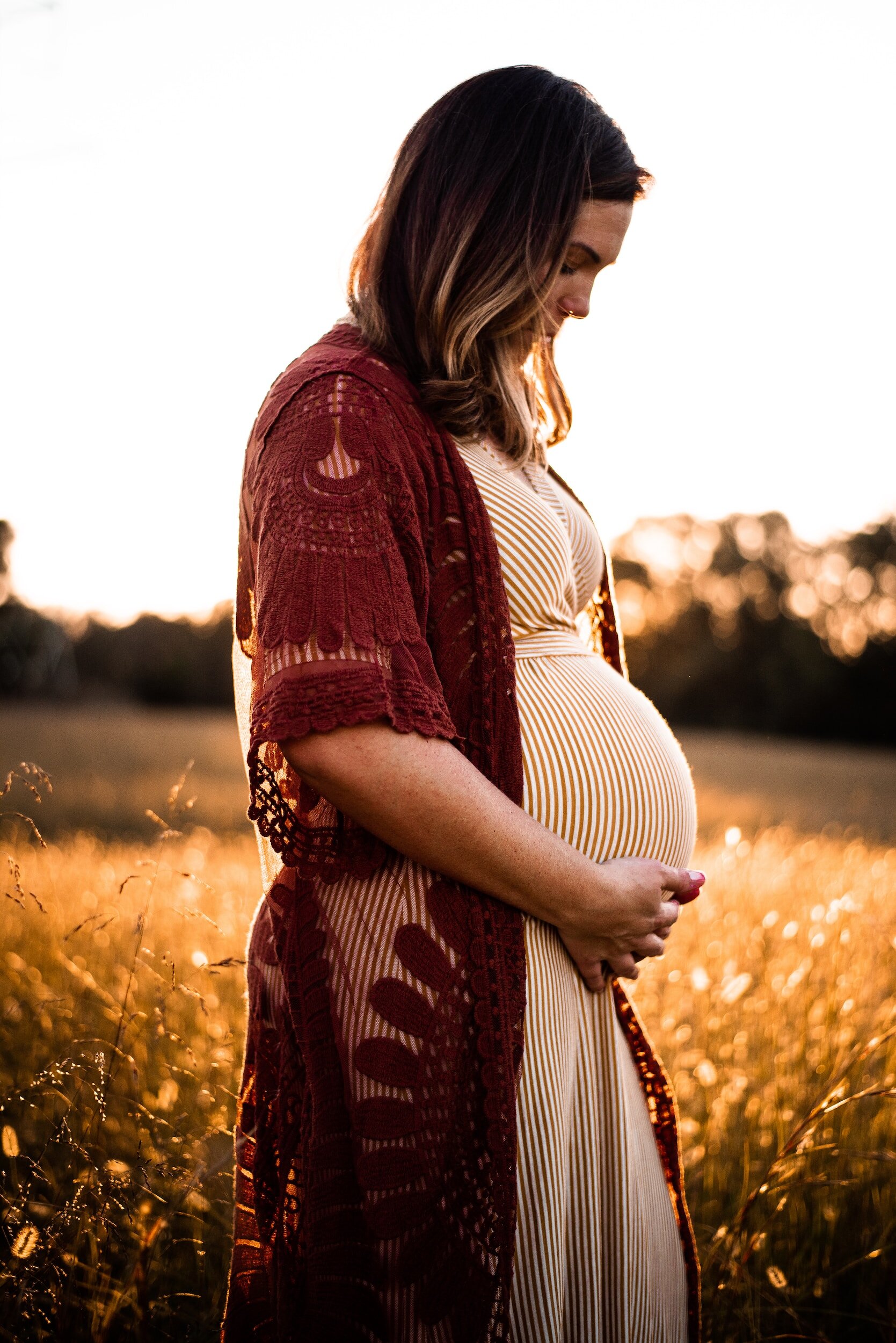 Pregnant woman standing in a field at sunset, holding her belly and wearing a striped dress with a maroon lace overlay.