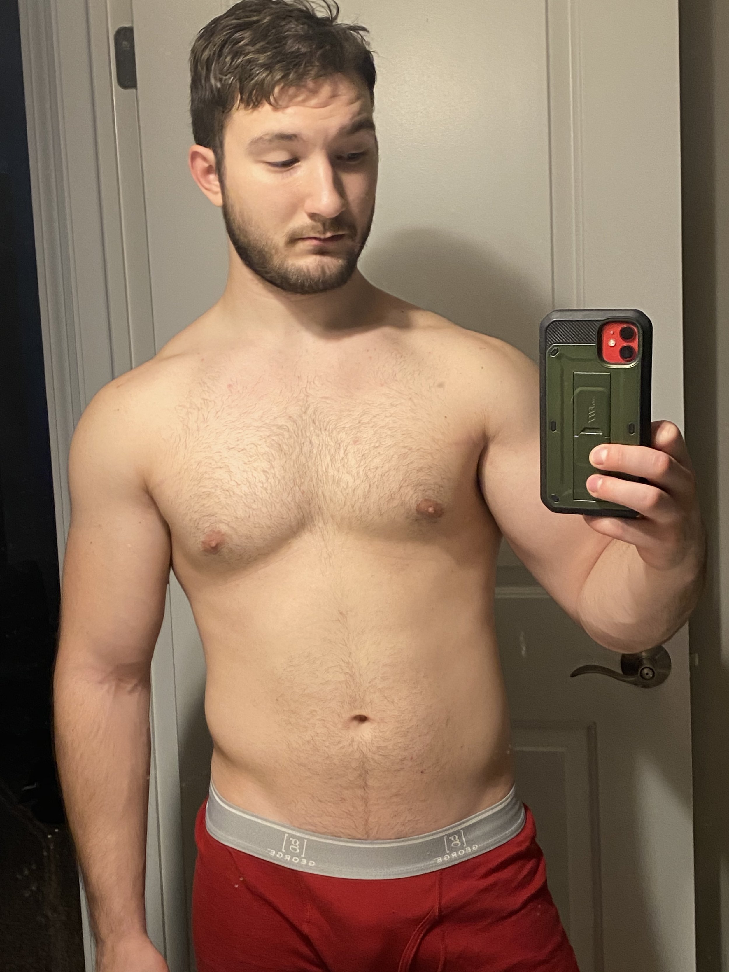 A shirtless young man with brown hair and a beard taking a selfie in front of a mirror. He is wearing red shorts with a gray waistband and is holding a green phone. The background shows a closed white door and part of a wooden door frame.