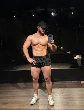 A muscular man taking a mirror selfie in a gym, wearing a black cap, glasses, black shorts, and white sneakers.