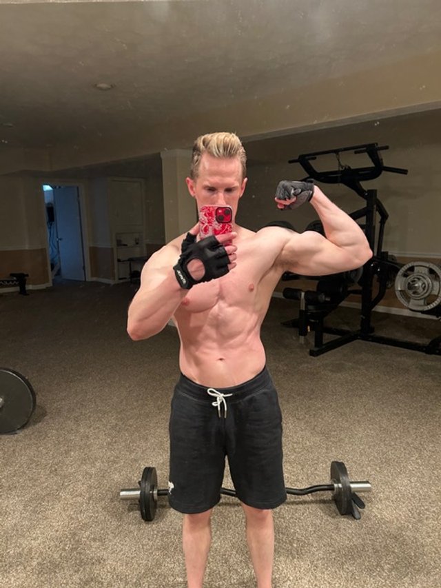 A young man with short blond hair taking a mirror selfie in a gym. He is shirtless, wearing black shorts, and black workout gloves, flexing his right bicep. Dumbbells and exercise equipment are visible in the background.