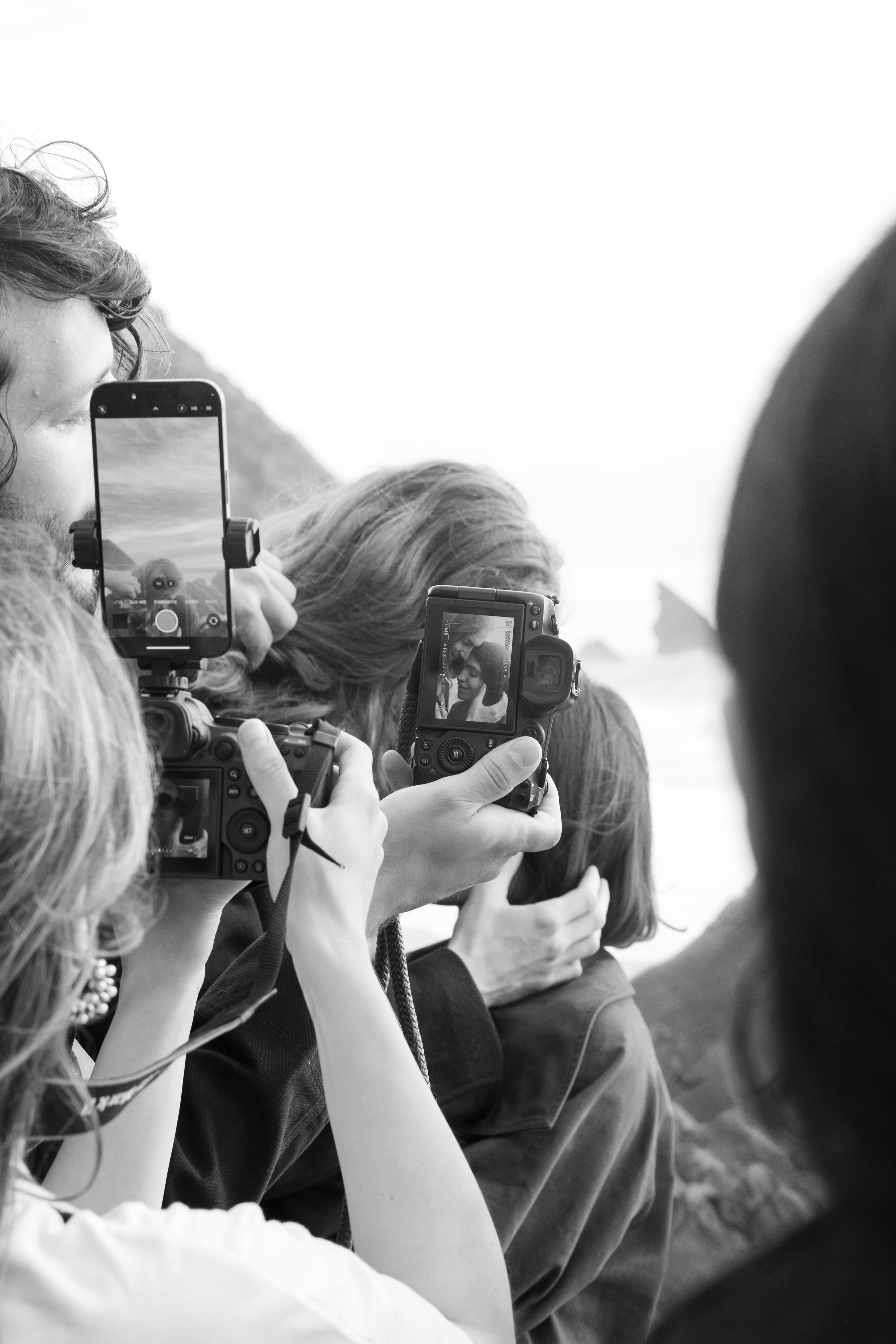 Multiple people holding cameras and smartphones, capturing a scene outdoors, with mountain and sky in the background.
