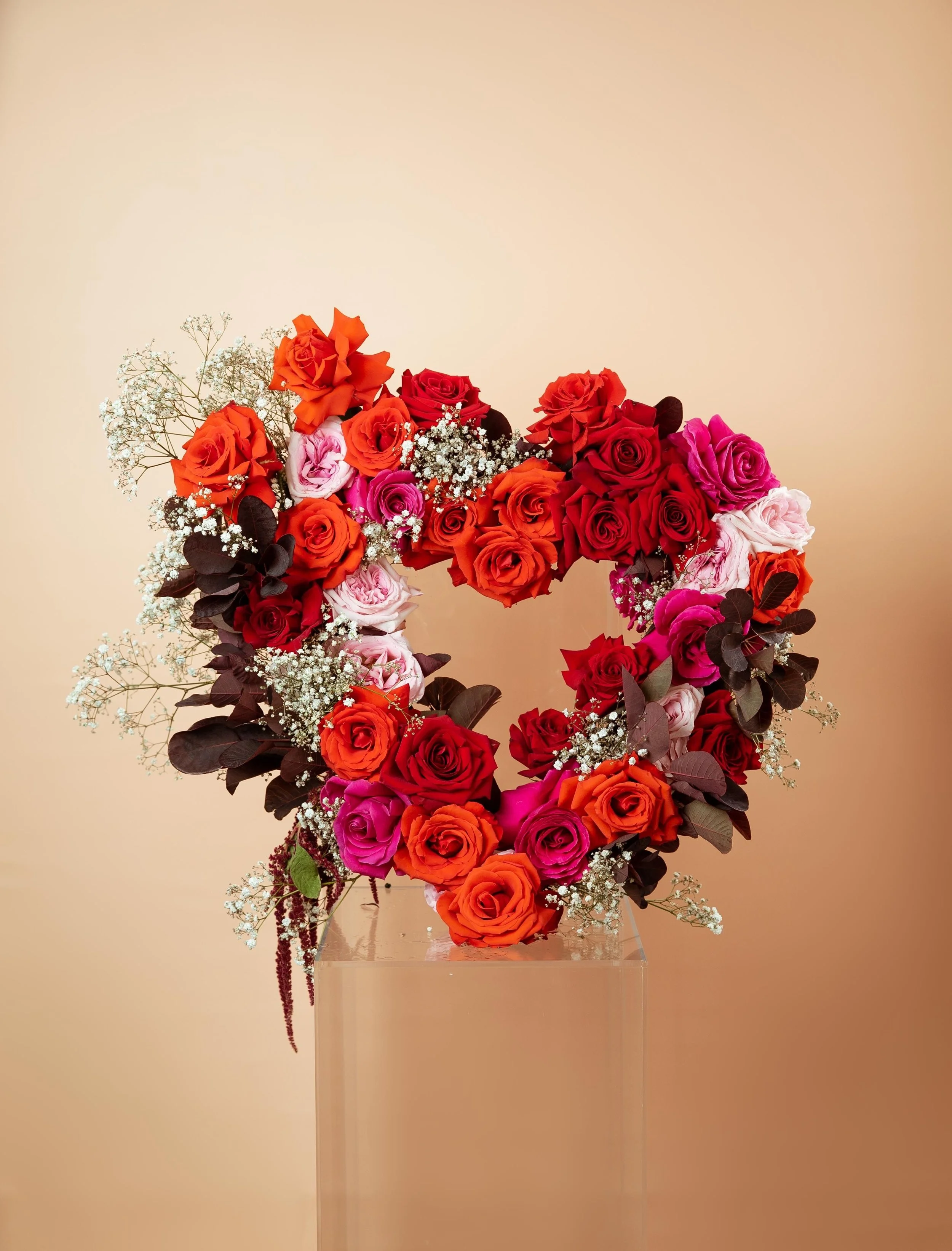 A heart-shaped floral arrangement made of red, pink, and orange roses, accented with white baby's breath and dark leaves, displayed on a clear stand against a beige background.