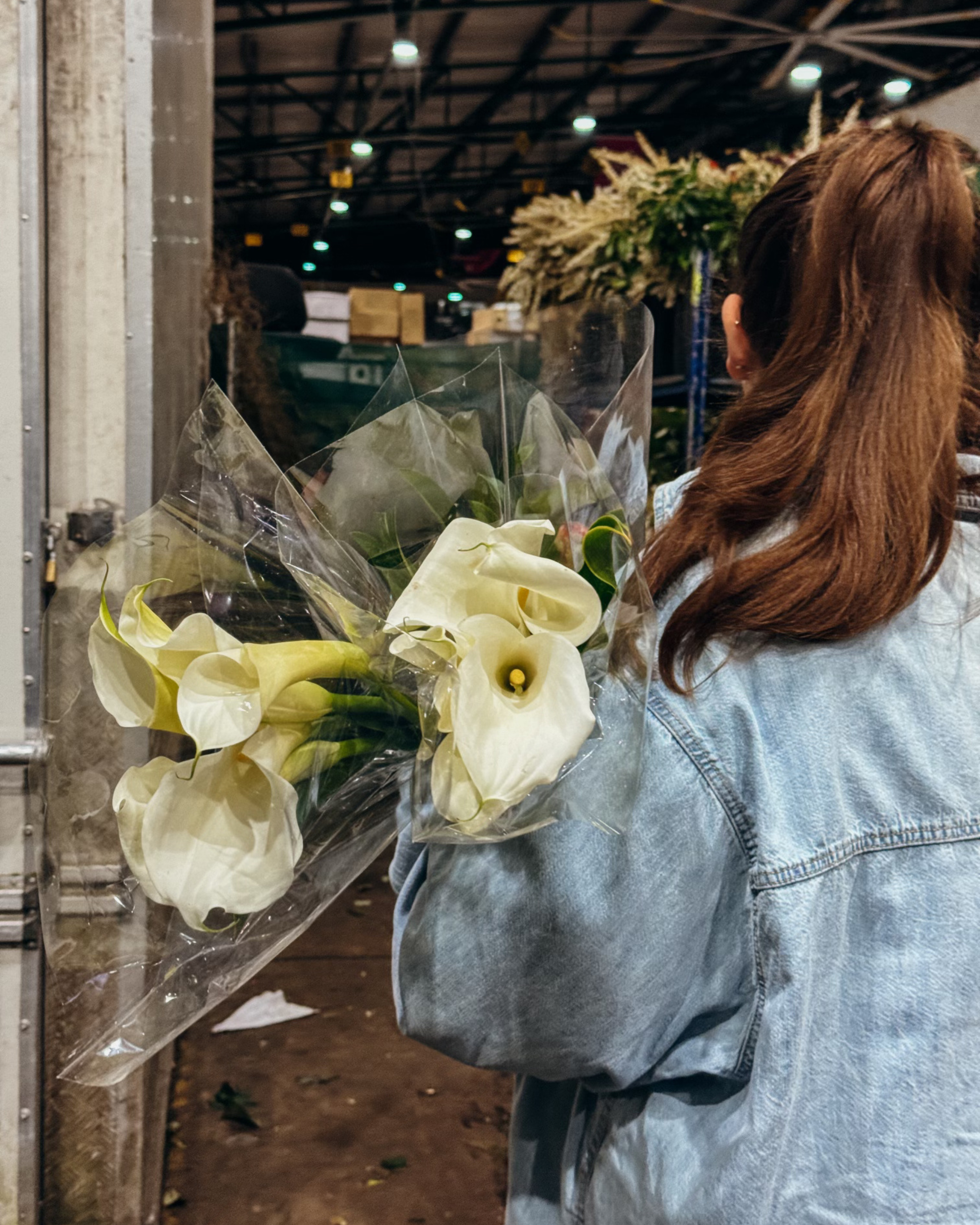 A woman holding a bouquet of white calla lilies wrapped in clear plastic at an indoor market.