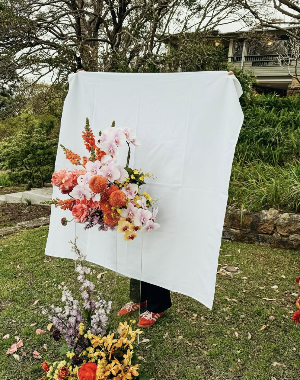 Person holding up a white backdrop with a large floral arrangement of pink, orange, yellow, and purple flowers in front of a garden with trees and a house in the background.