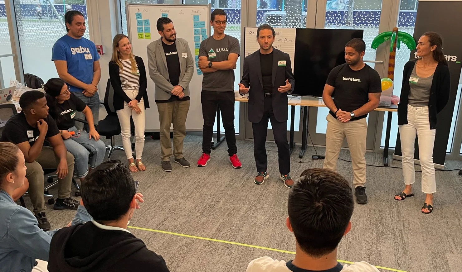 Group of people in a meeting room standing and discussing, with whiteboards and notes in the background.