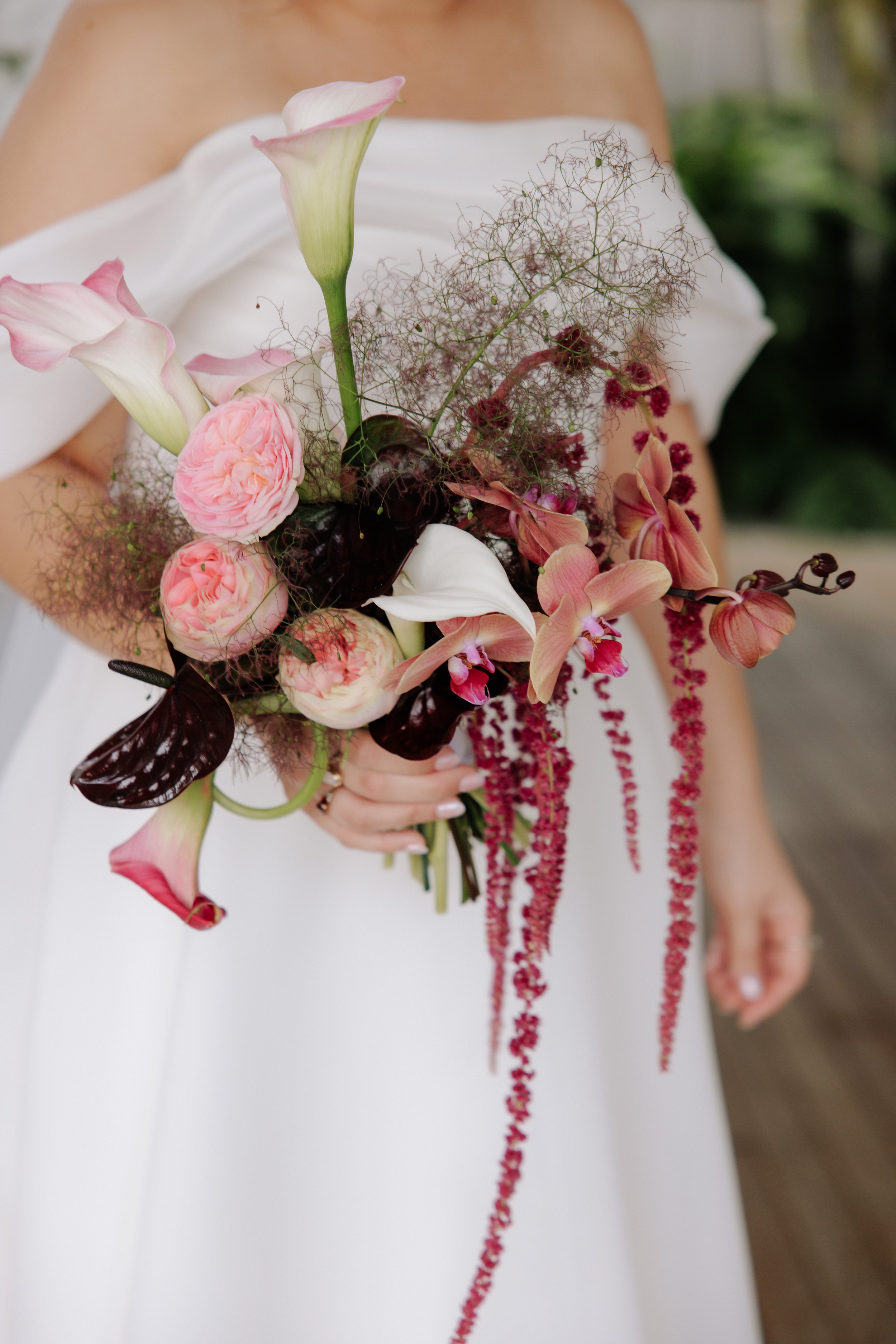 A woman in a white dress holding a bouquet of pink and white flowers, including calla lilies, roses, and orchids, with pink bead strands hanging down.