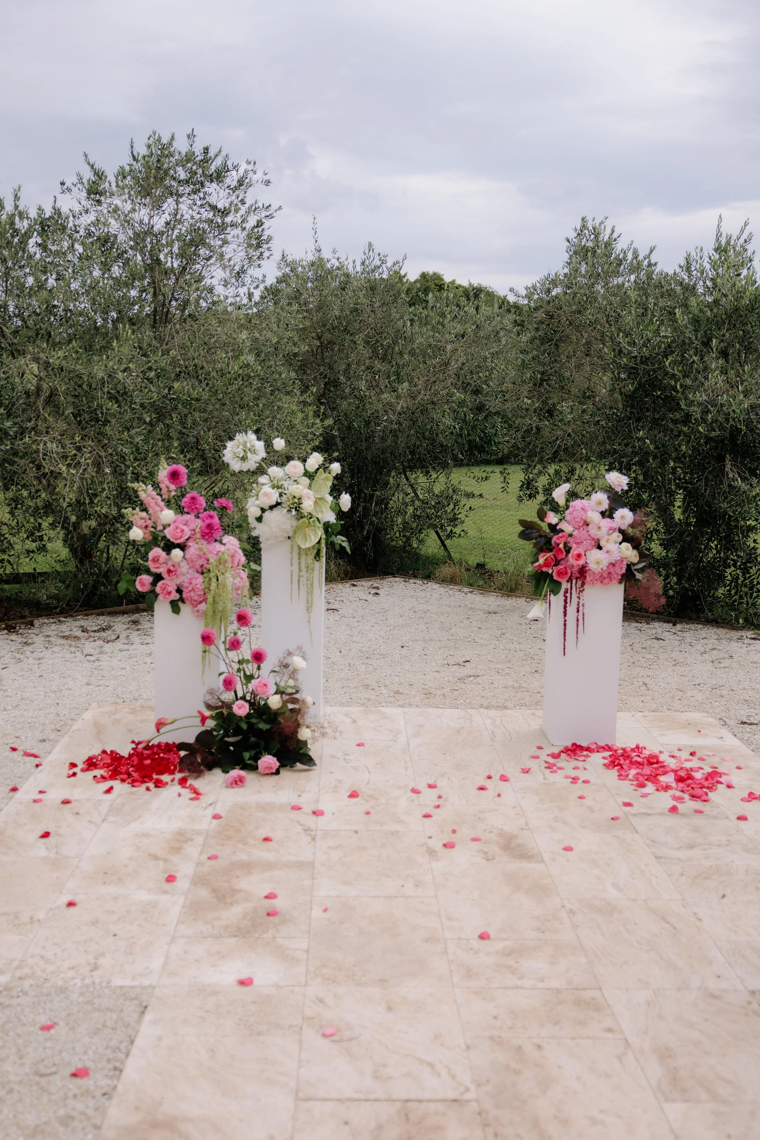 Outdoor wedding altar decorated with pink and white flowers, red rose petals scattered on the ground, set against a backdrop of green trees and a cloudy sky.