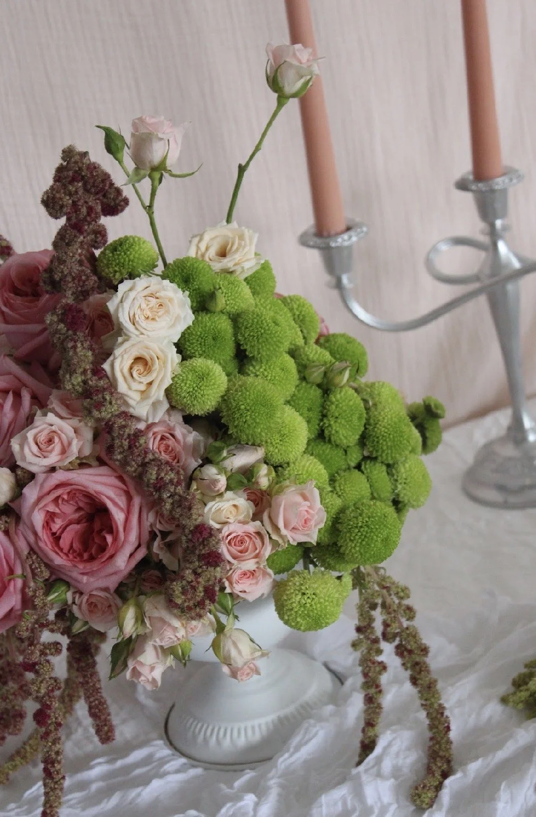 A floral arrangement with pink roses, white roses, and green pom-pom flowers in a white vase, with a pink candleholder and candles in the background.
