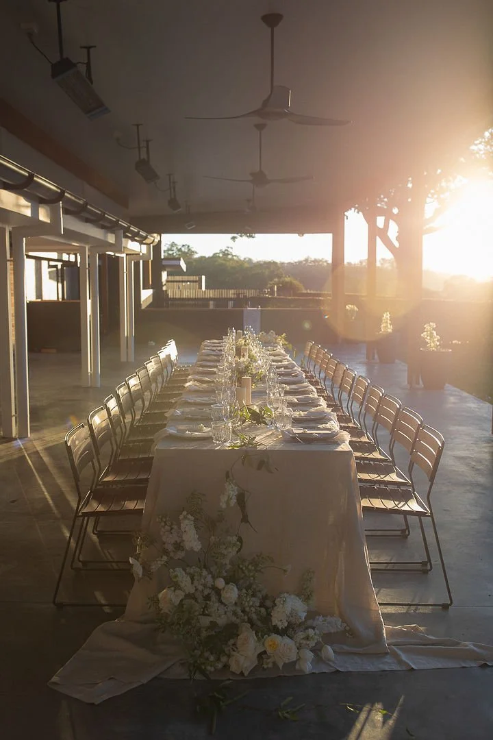 Long dining table set for a formal event with white tablecloth, candles, glassware, and floral centerpieces, outdoors at sunset with surrounding trees and potted plants.