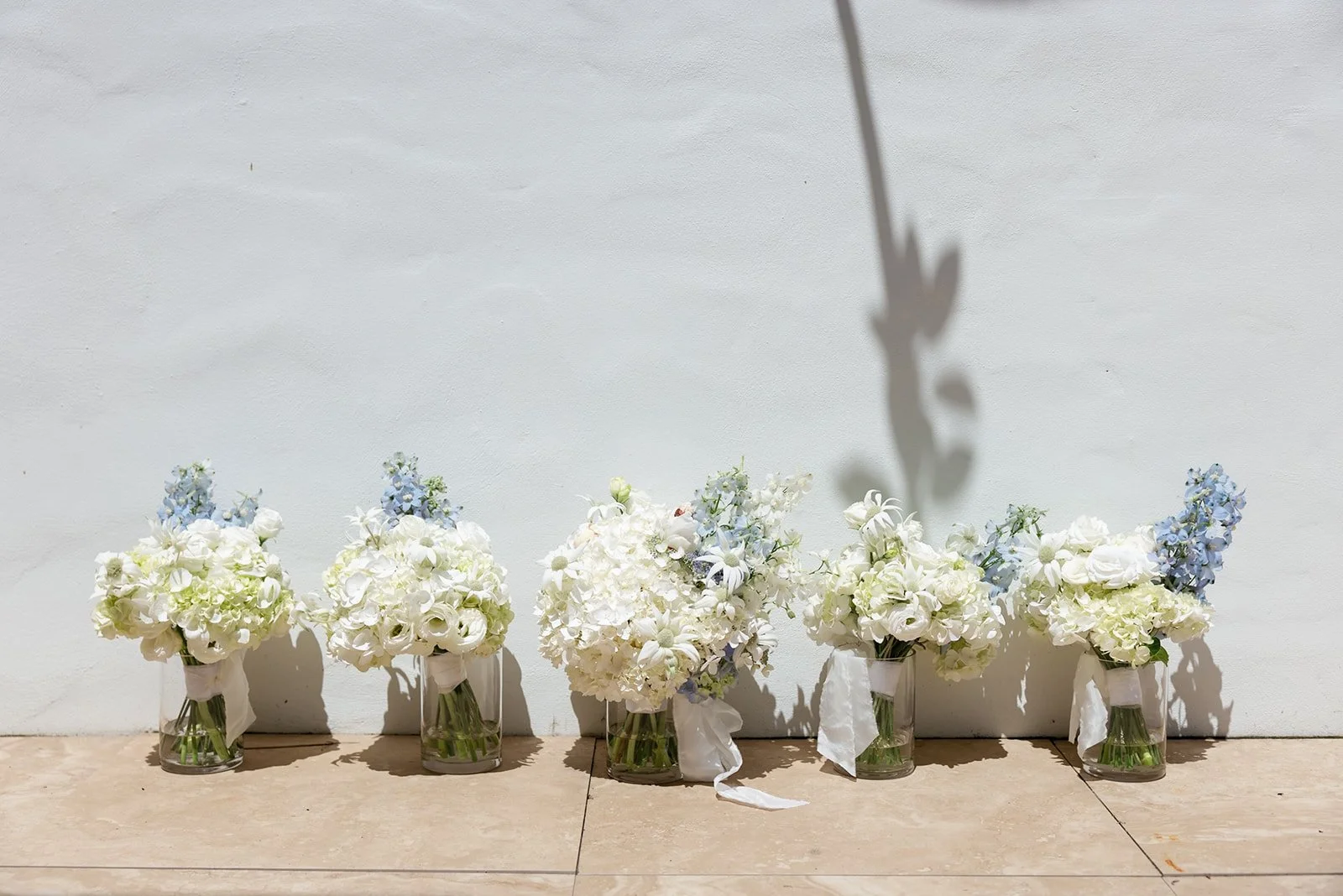 Six vases filled with white and pale blue flowers placed against a white wall, with a shadow of a single flower cast on the wall.