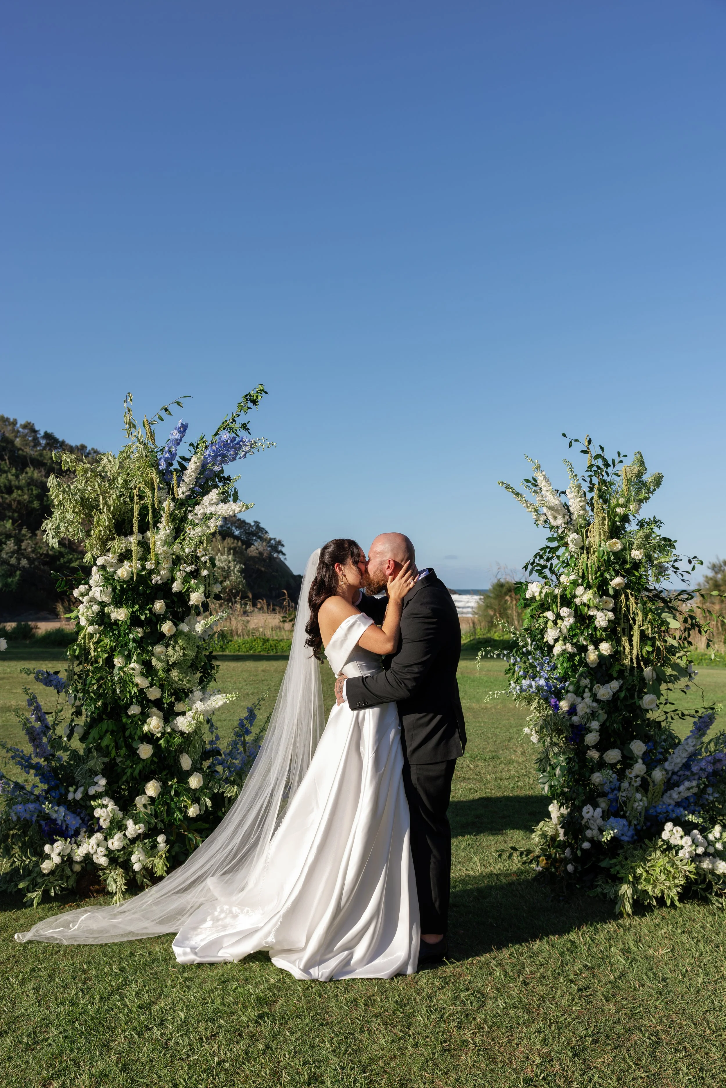 A bride and groom kiss under a floral arch at an outdoor wedding ceremony near the ocean on a sunny day.