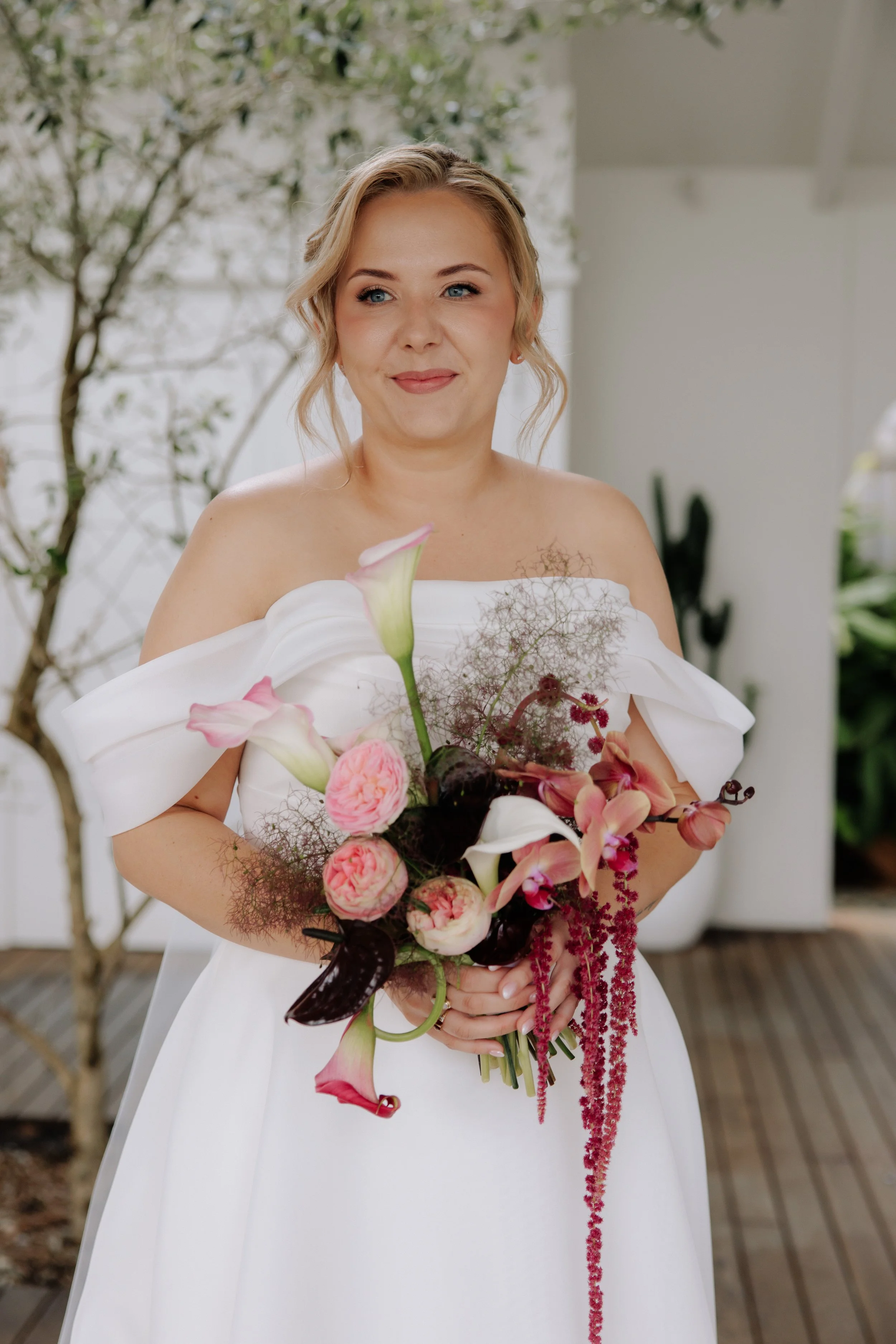 A woman in a white dress holding a bouquet of pink, white, and red flowers, standing on a wooden deck with greenery in the background.