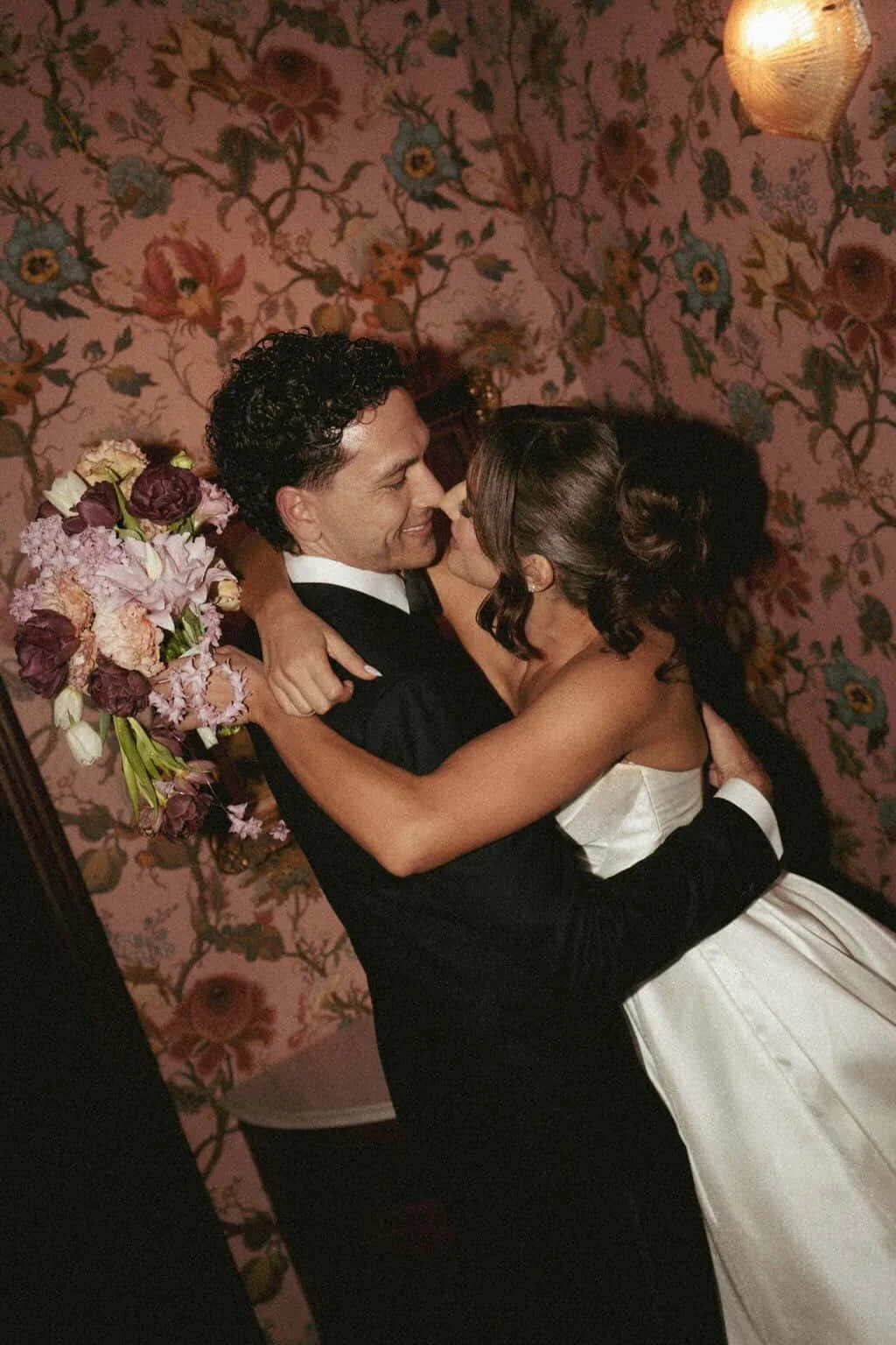 A couple dancing closely and smiling, with the woman holding a bouquet of flowers, in a room with floral wallpaper and warm lighting.