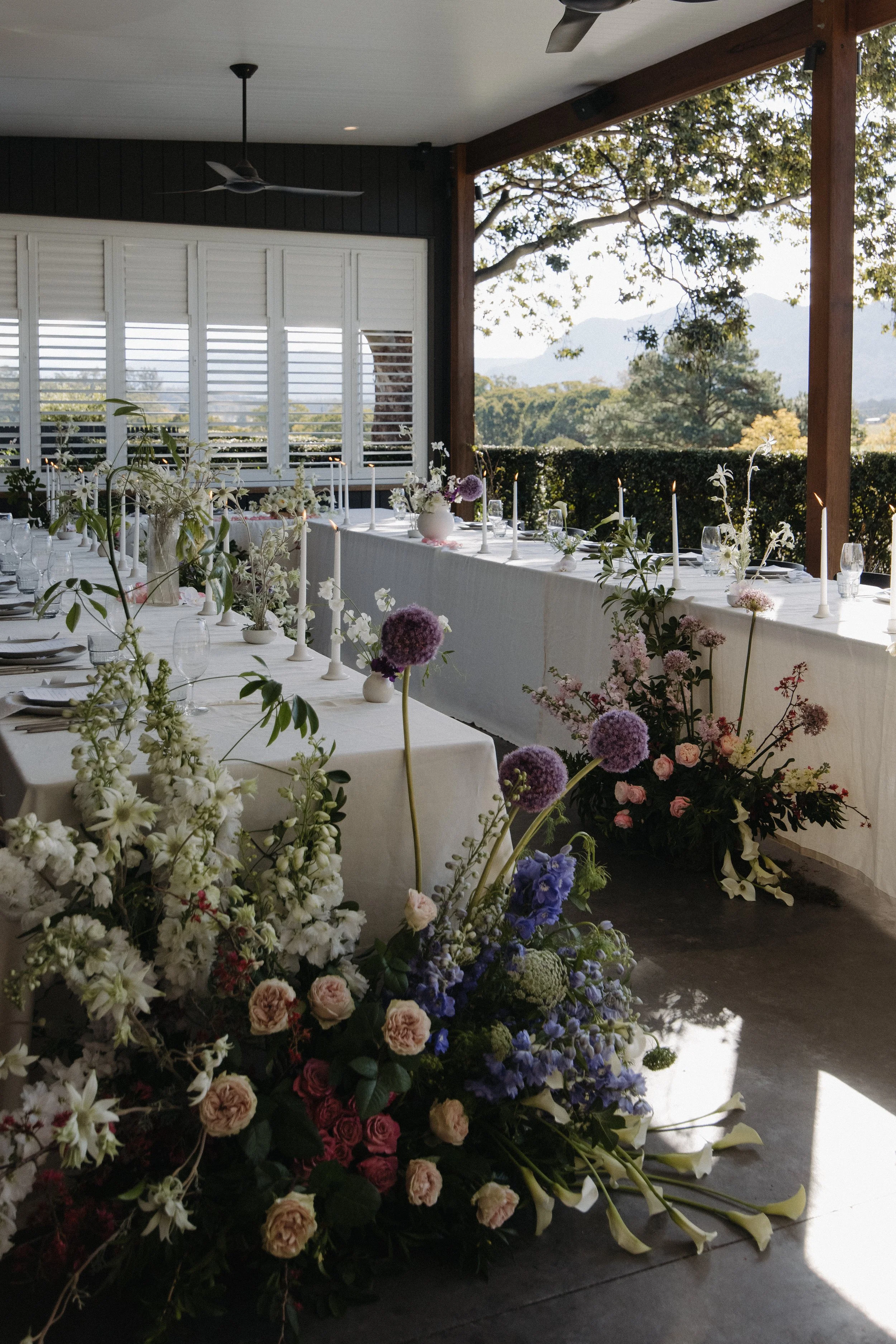 A long banquet table decorated with a white tablecloth and floral arrangements, with candles and glassware, set up in an outdoor covered patio area overlooking trees and mountains in the distance.