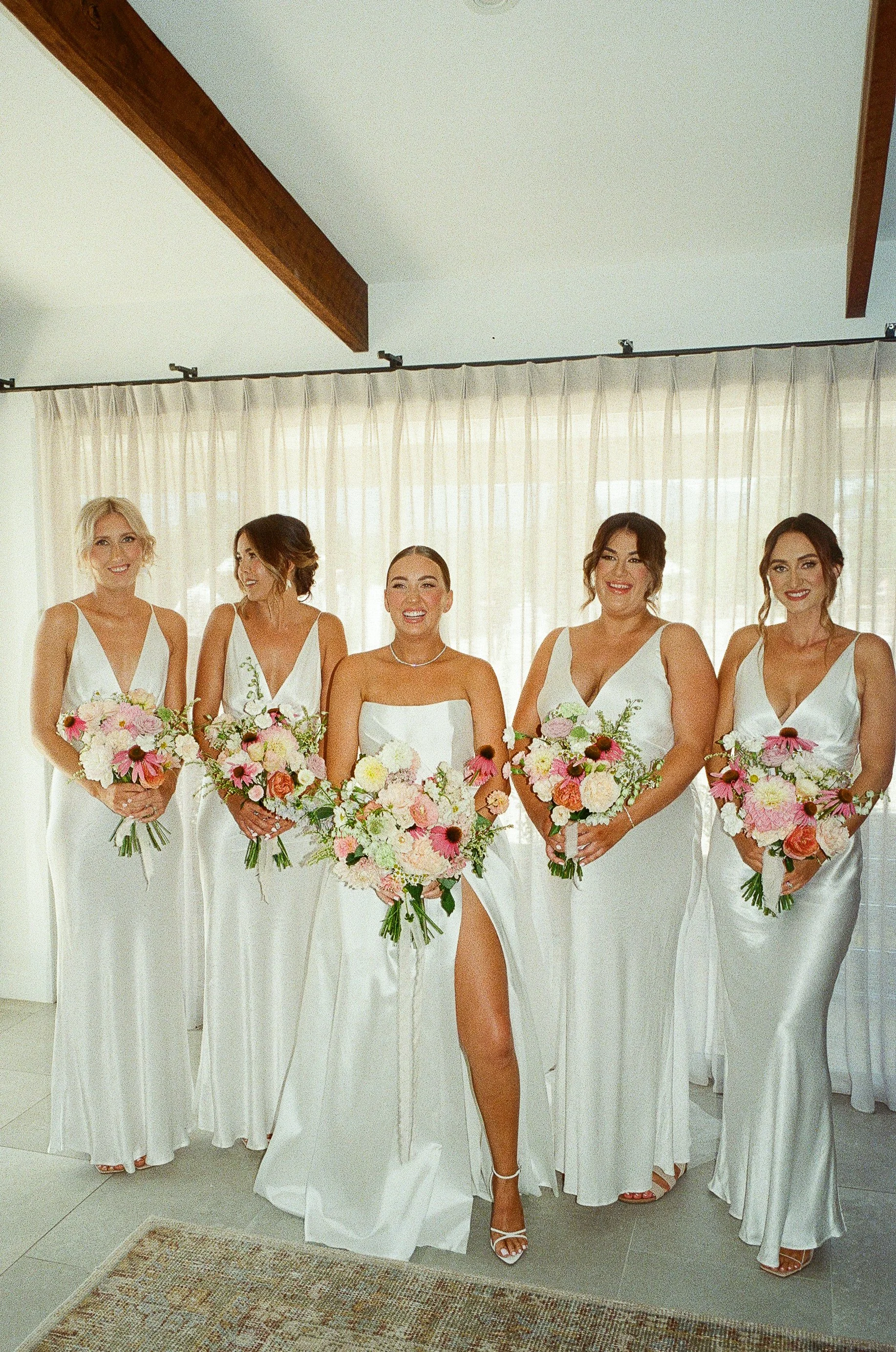 A group of five women dressed in white gowns holding bouquets of pink, white, and green flowers, standing inside a room with white curtains and wooden beams.