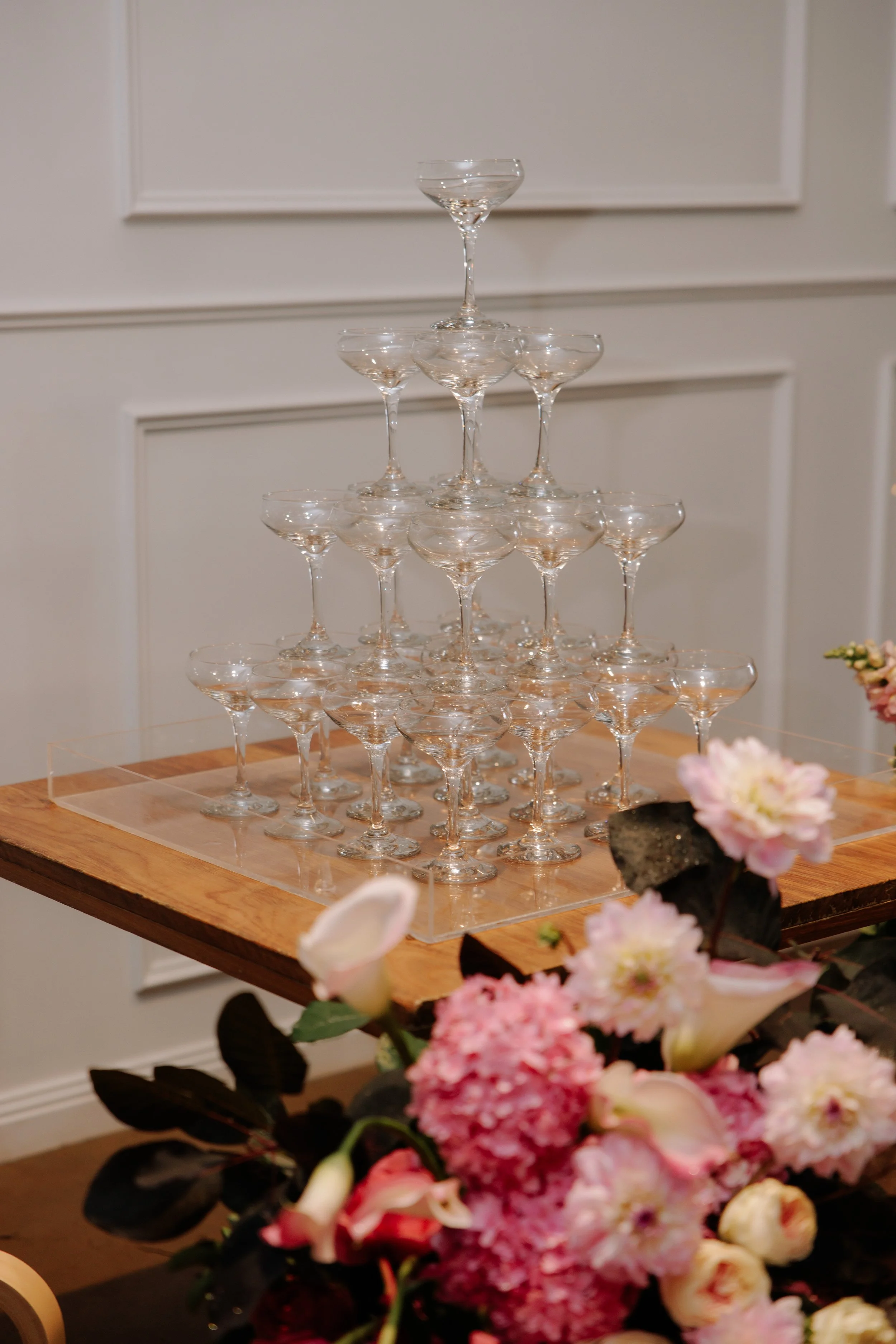 A pyramid of empty champagne coupes arranged on a wooden table with a floral centerpiece in the foreground.