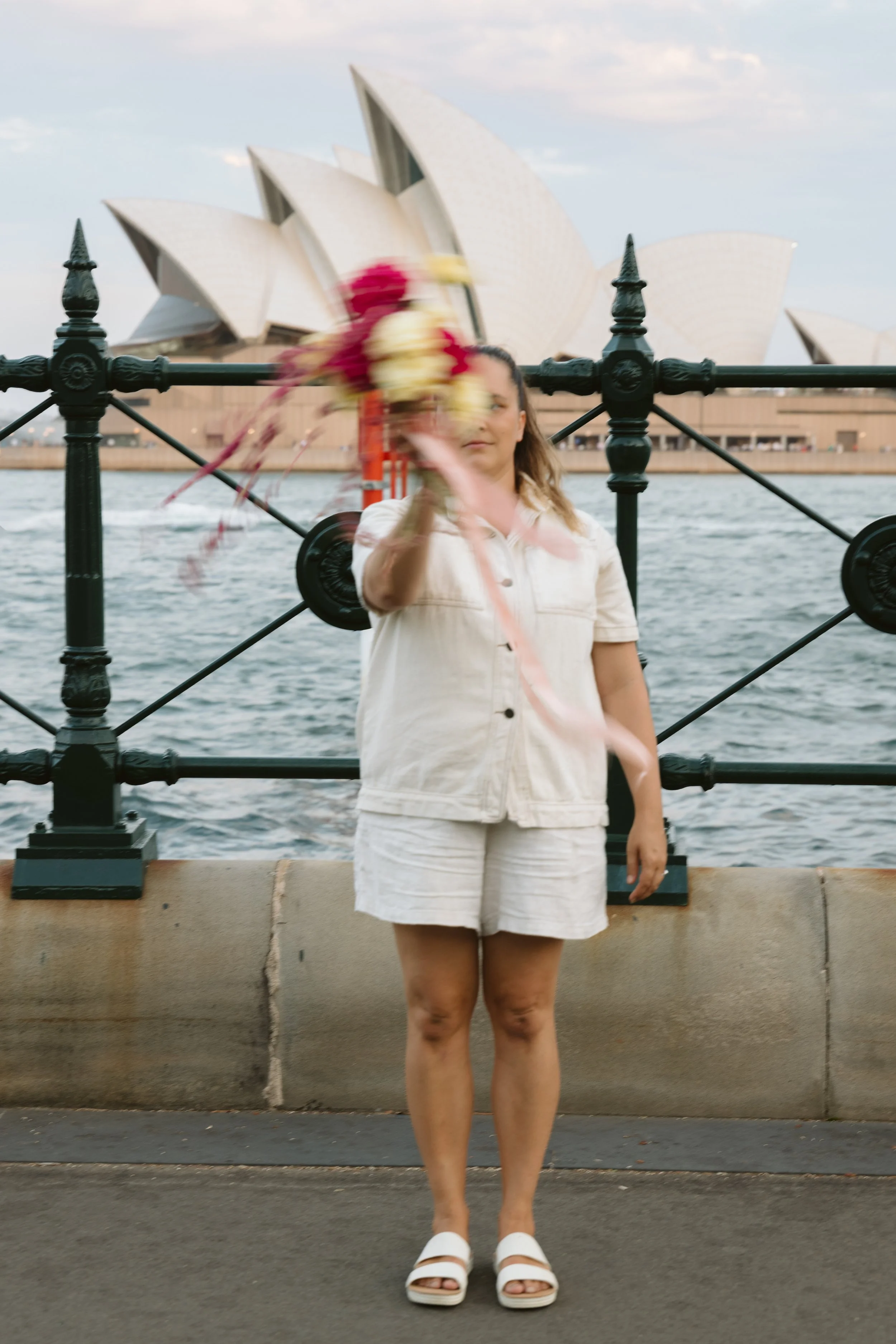 A woman stands on a waterfront promenade with the Sydney Opera House in the background, holding a bouquet of flowers blurred in motion in front of her face.