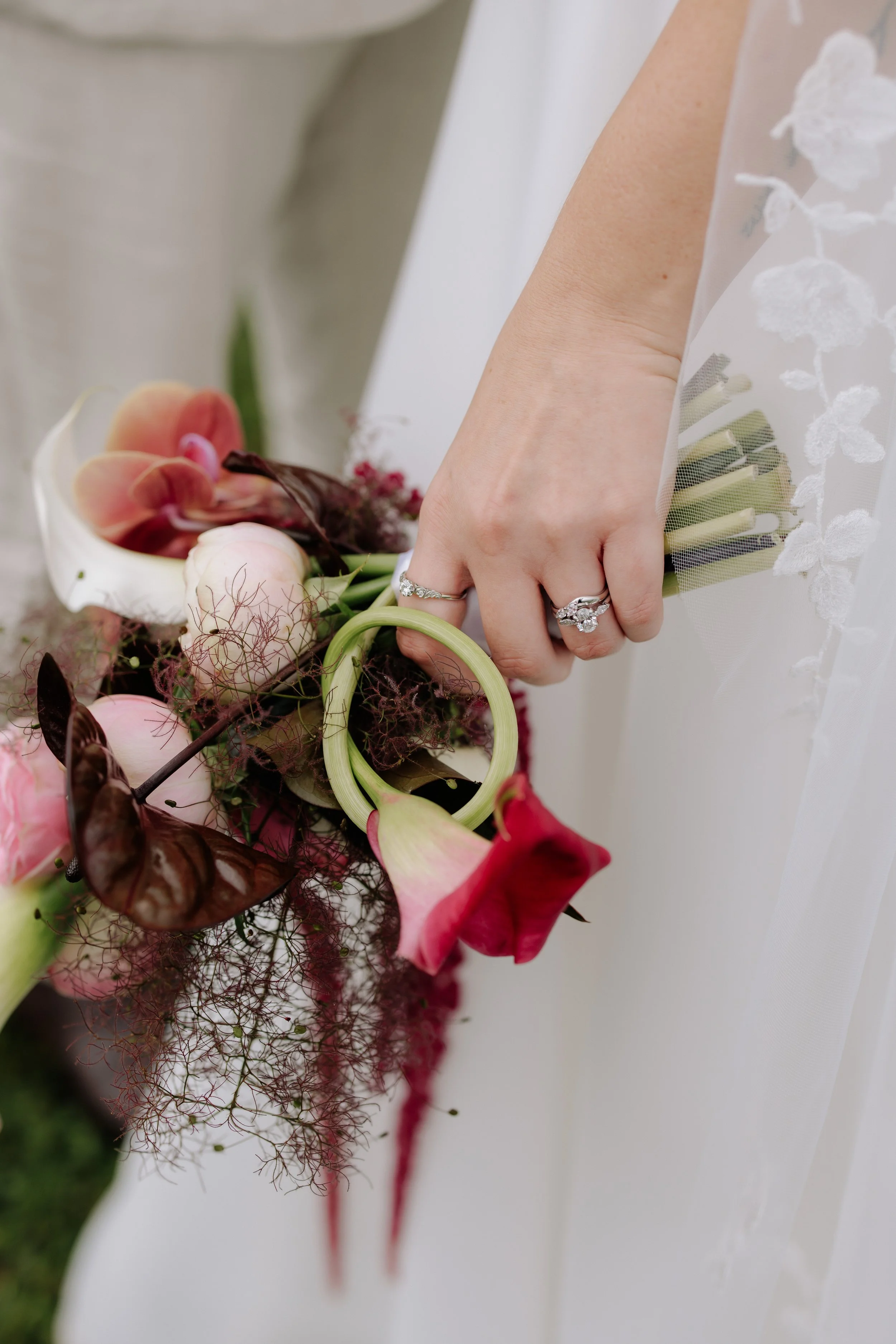 Close-up of a person's hand holding a bouquet of various flowers, including calla lilies, roses, and orchids, with a lace wedding dress visible.