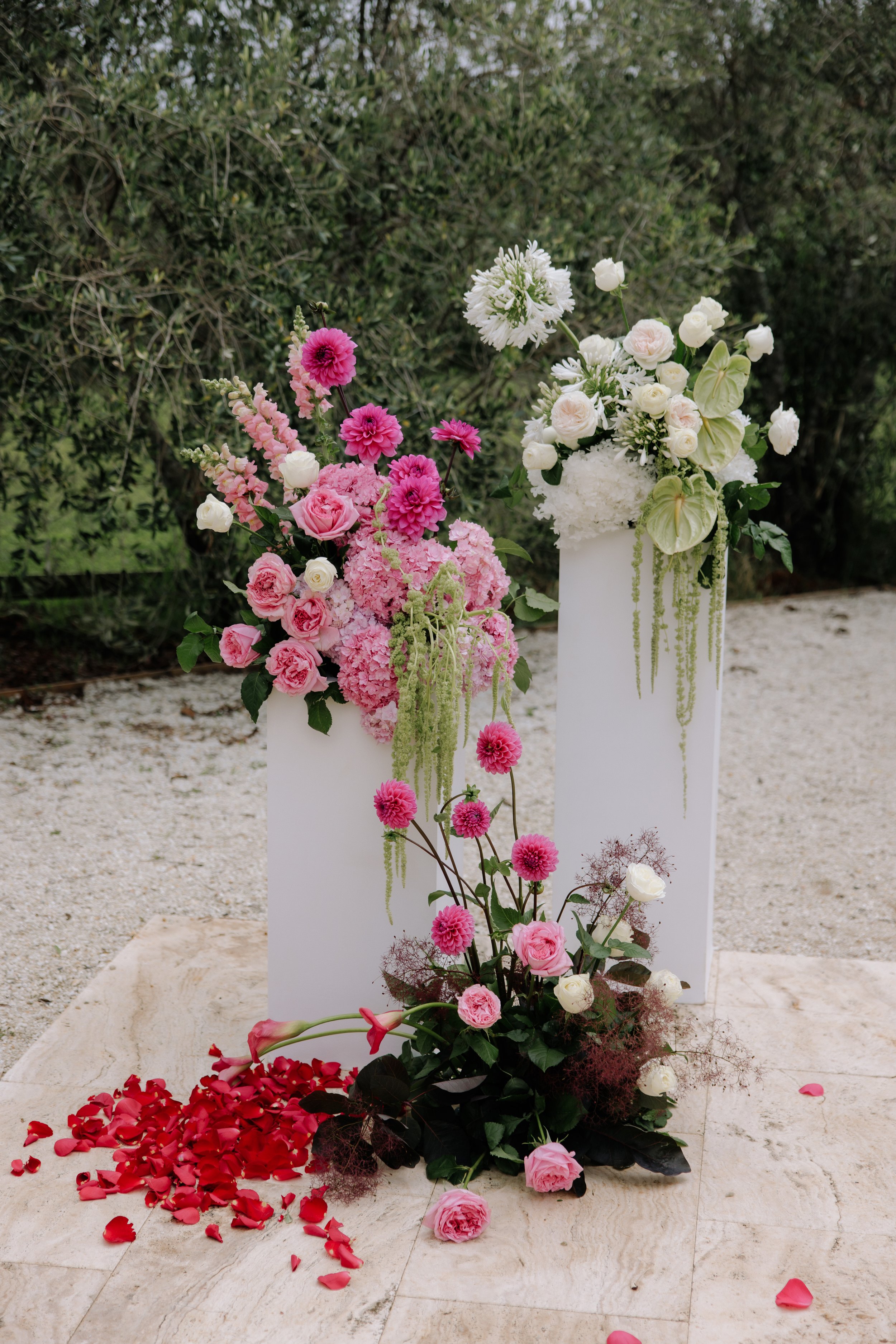 Elegant outdoor display of pink and white floral arrangements on white pedestals, with scattered rose petals at the base, set against a background of greenery.