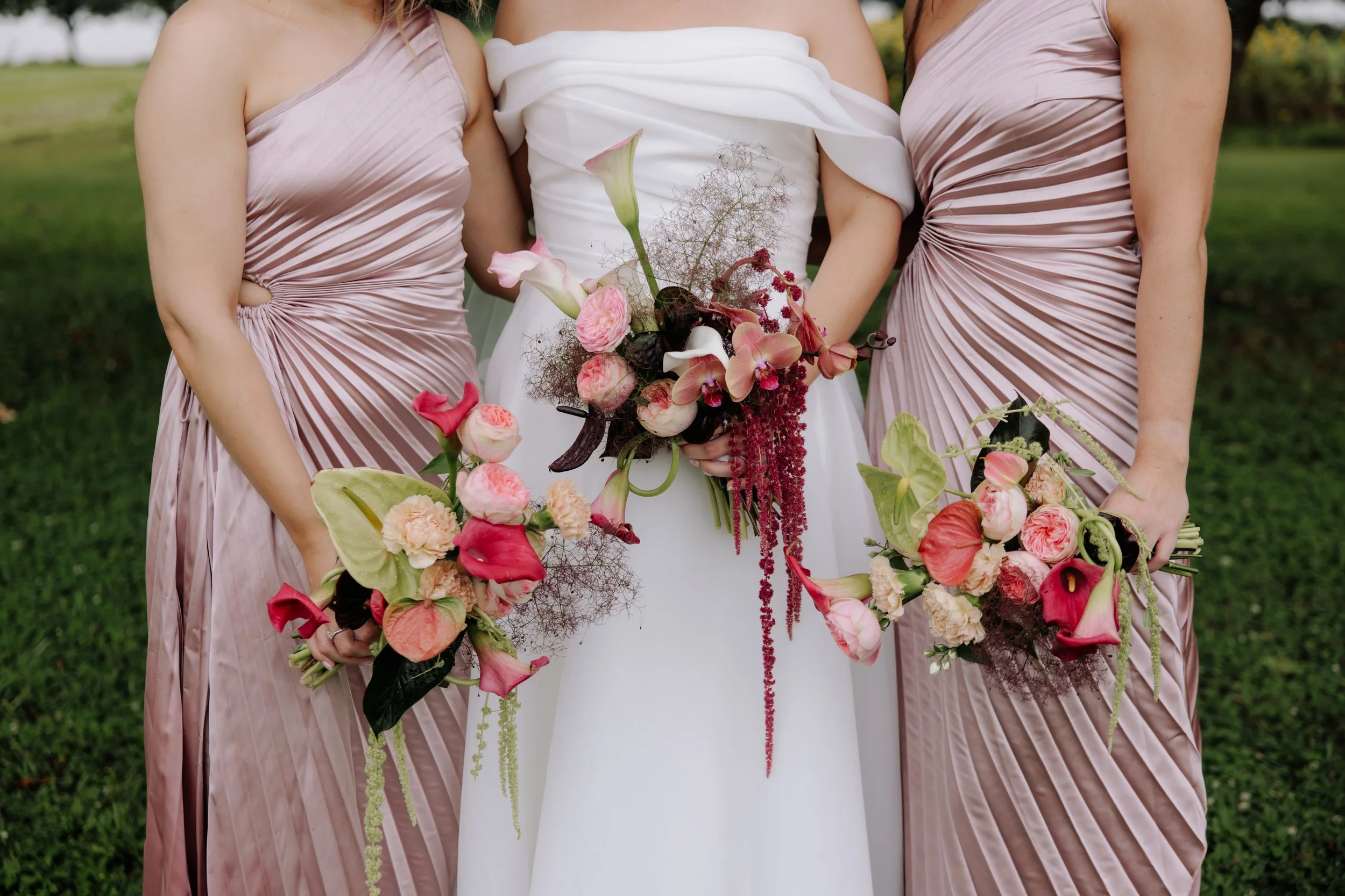 Close-up of three women, with the middle woman wearing a white gown and holding a bouquet of flowers, while the other two women wear matching satin dresses in light pink or mauve, standing on a grassy area outdoors.