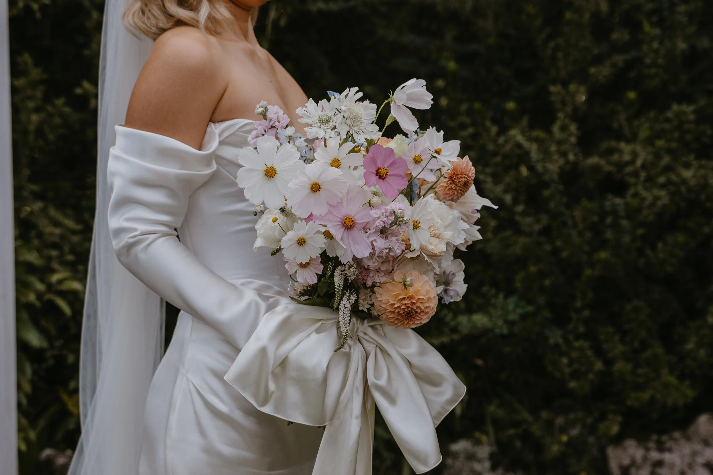 A bride in a satin white wedding dress holding a bouquet of white, pink, and peach flowers against a background of greenery.