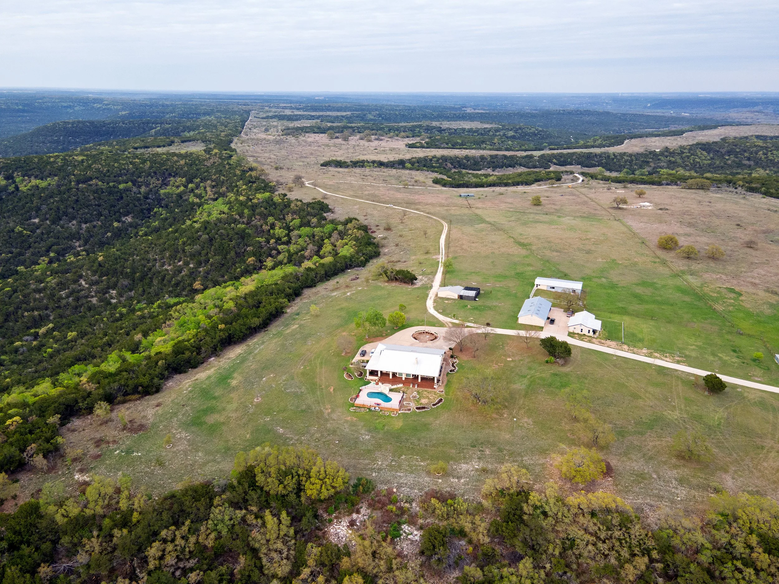 Aerial view of a rural landscape with houses, a swimming pool, trees, open fields, and winding roads, surrounded by forested areas.