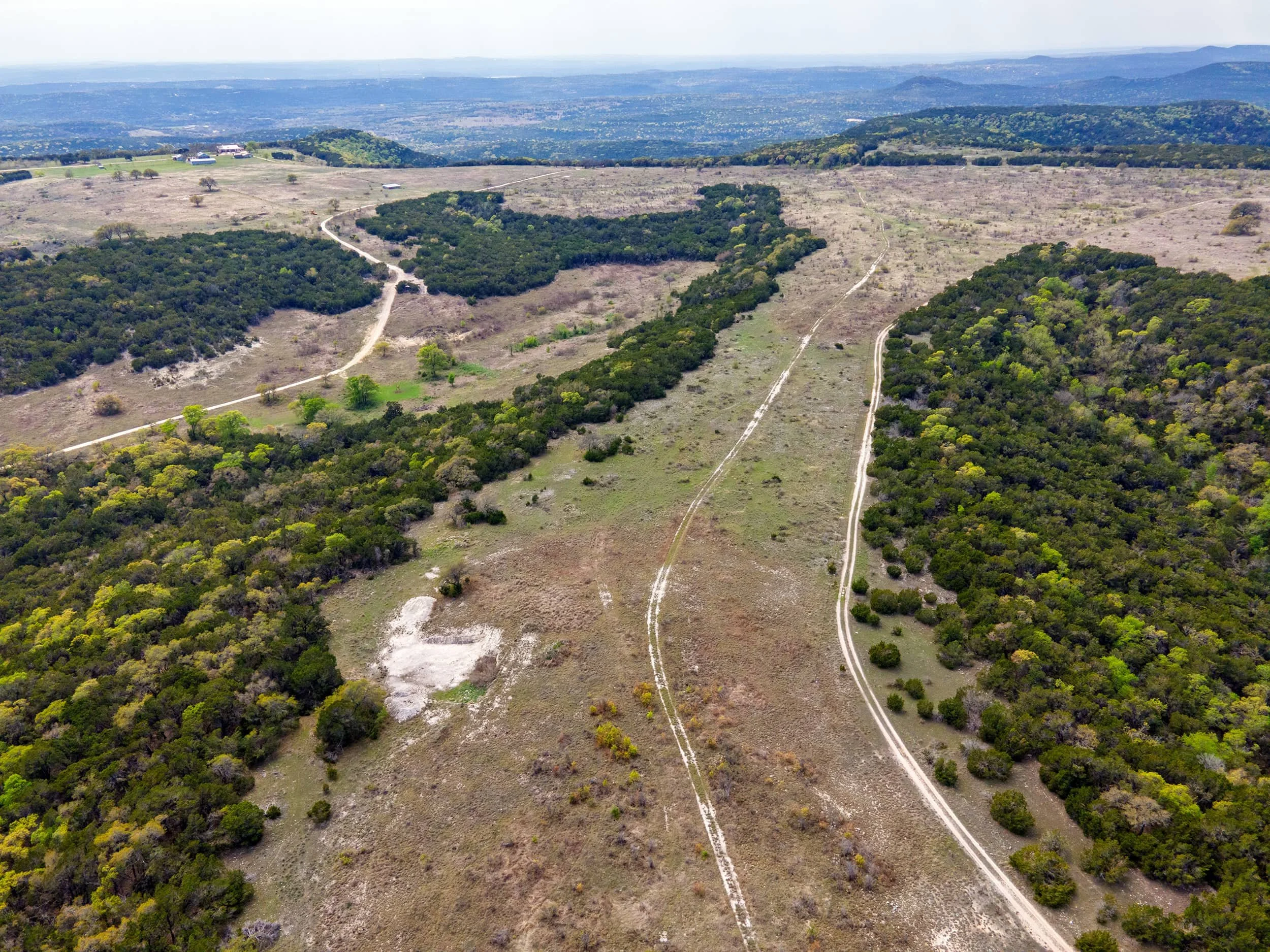 Aerial view of a hilly landscape with dirt roads, patches of green trees, and open fields under a cloudy sky.