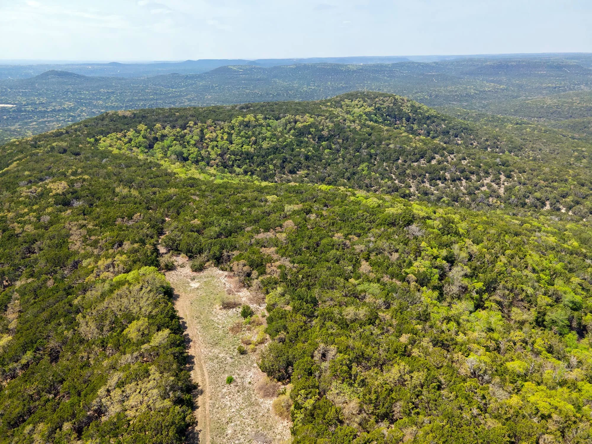 Aerial view of green rolling hills and dense forest under a cloudy sky.