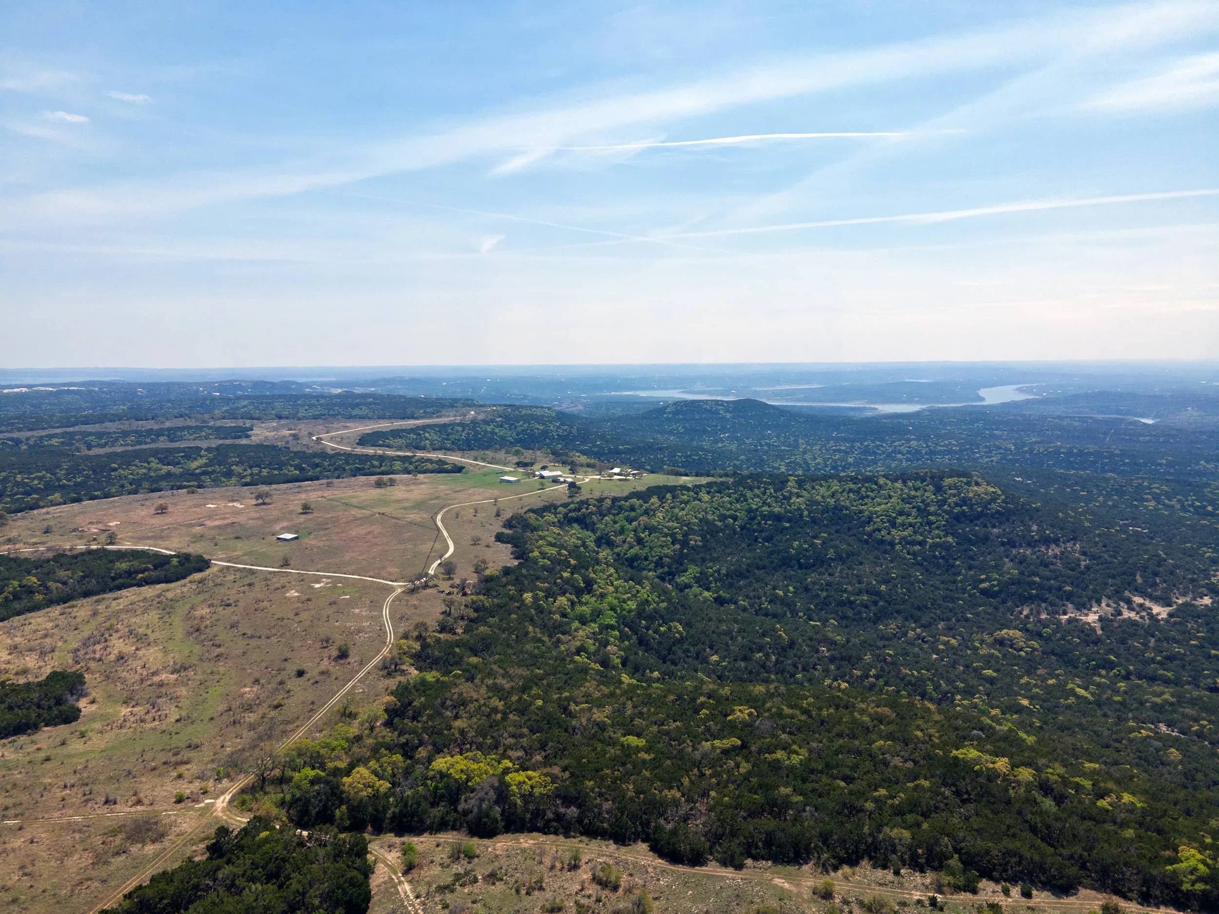 Aerial view of a rural landscape with hills, trees, a winding road, a few buildings, and a large body of water in the distance under a partly cloudy sky.