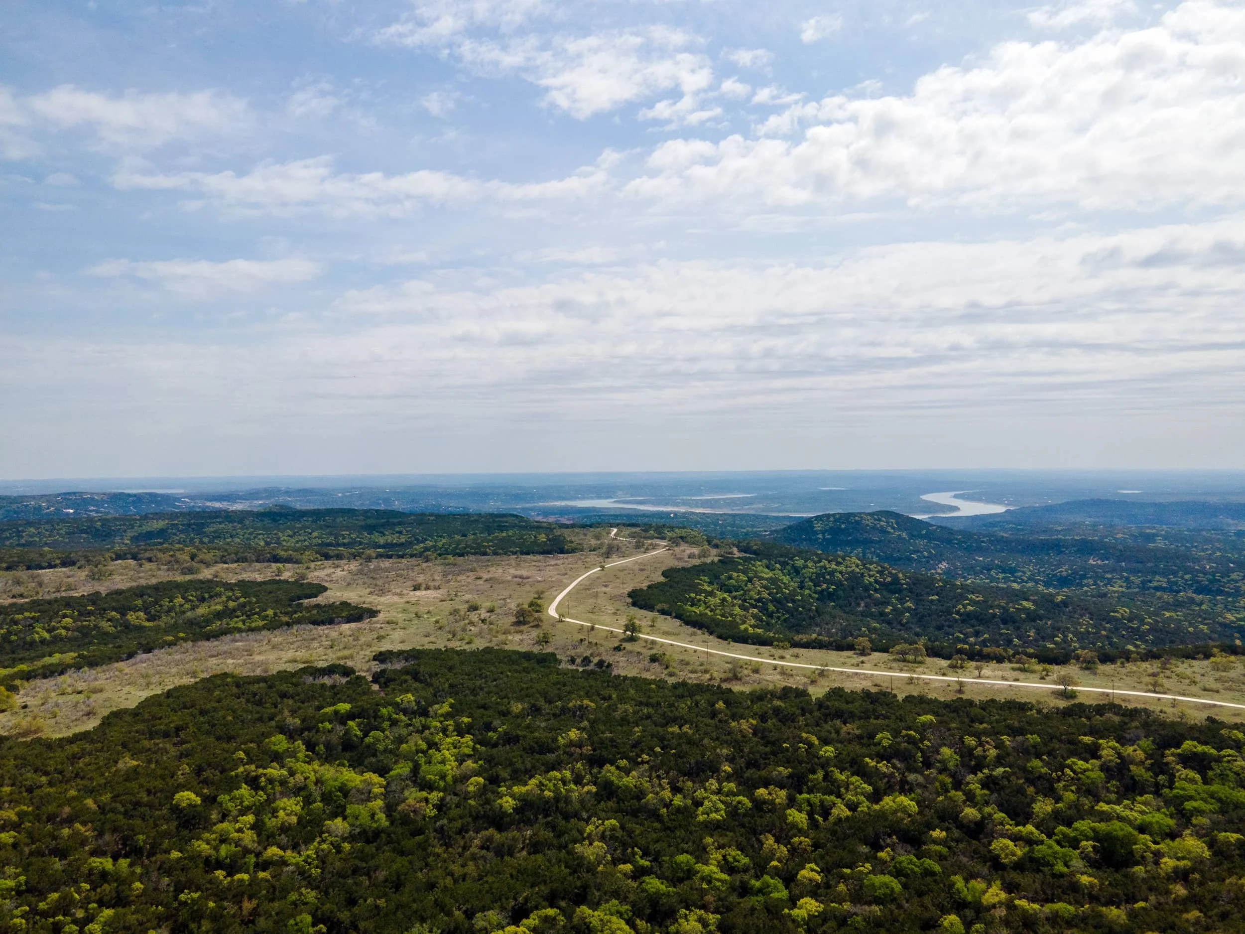 Vast landscape view with a winding road through forested hills, distant water bodies, and a bright blue sky with scattered clouds.