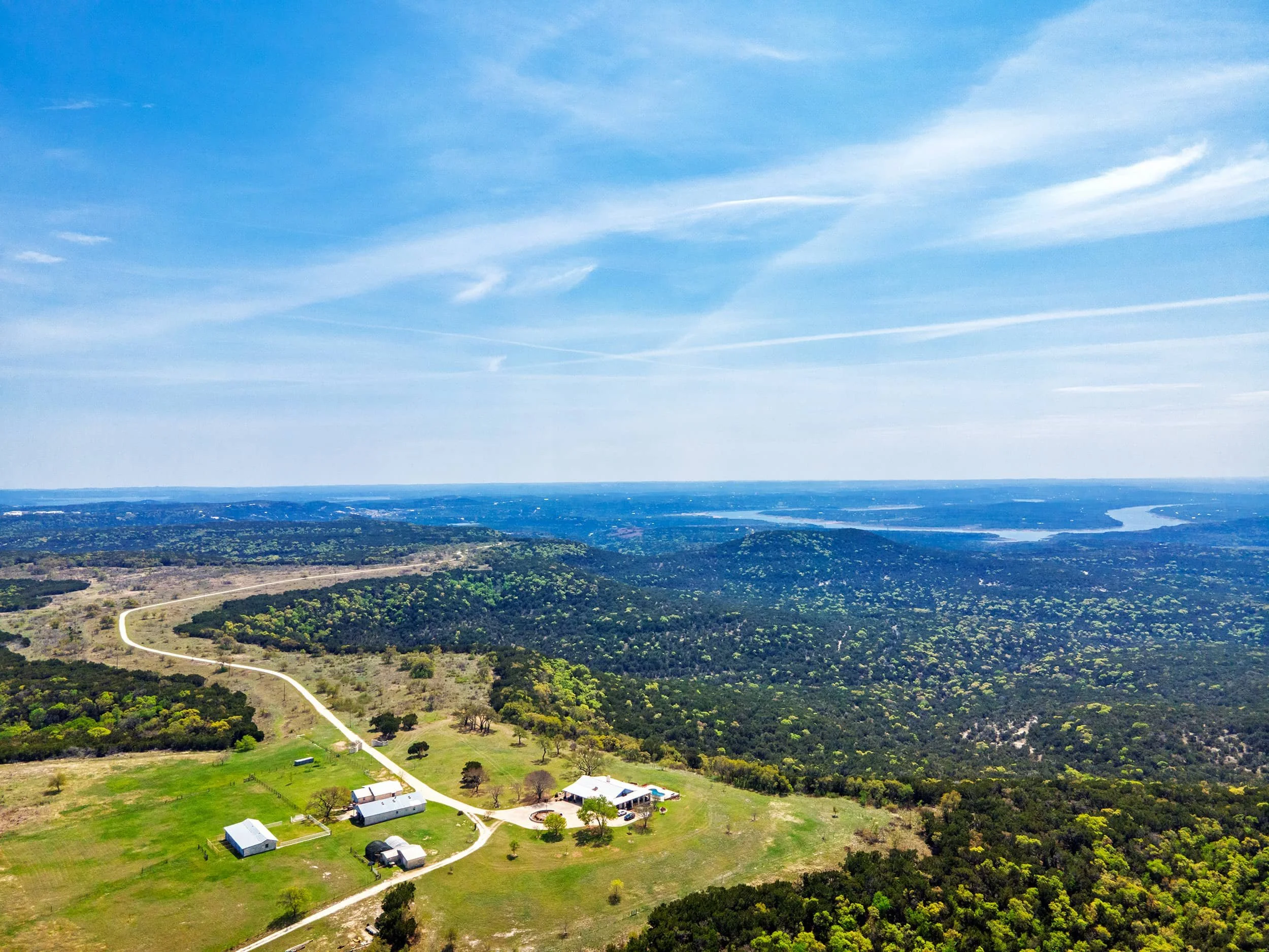 Aerial view of a rural landscape with a few buildings, roads, green fields, dense trees, a river, and a vast blue sky with scattered clouds.
