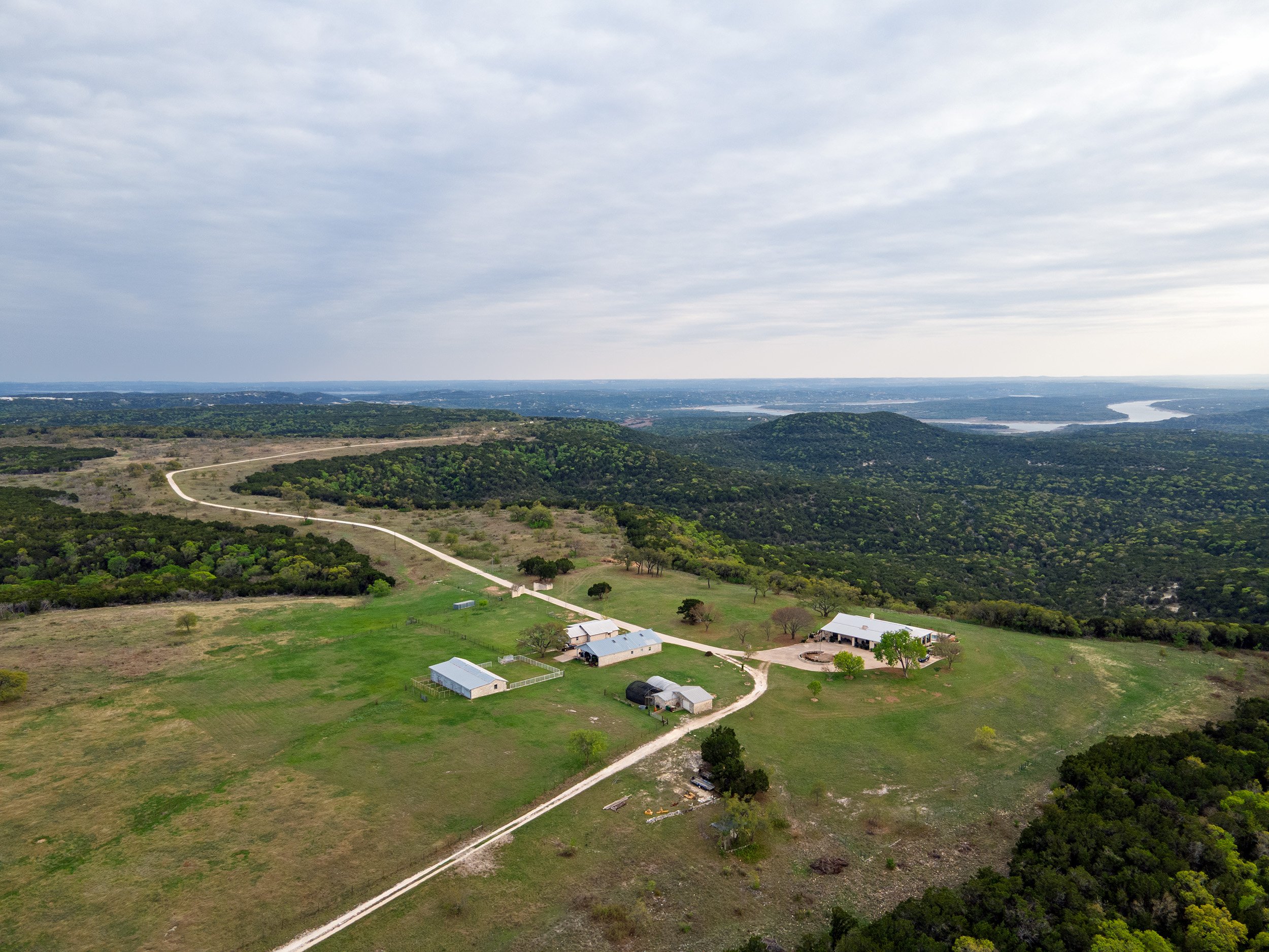 Aerial view of a rural landscape with green fields, a winding dirt road, farm buildings, trees, hills, a river, and cloudy sky.