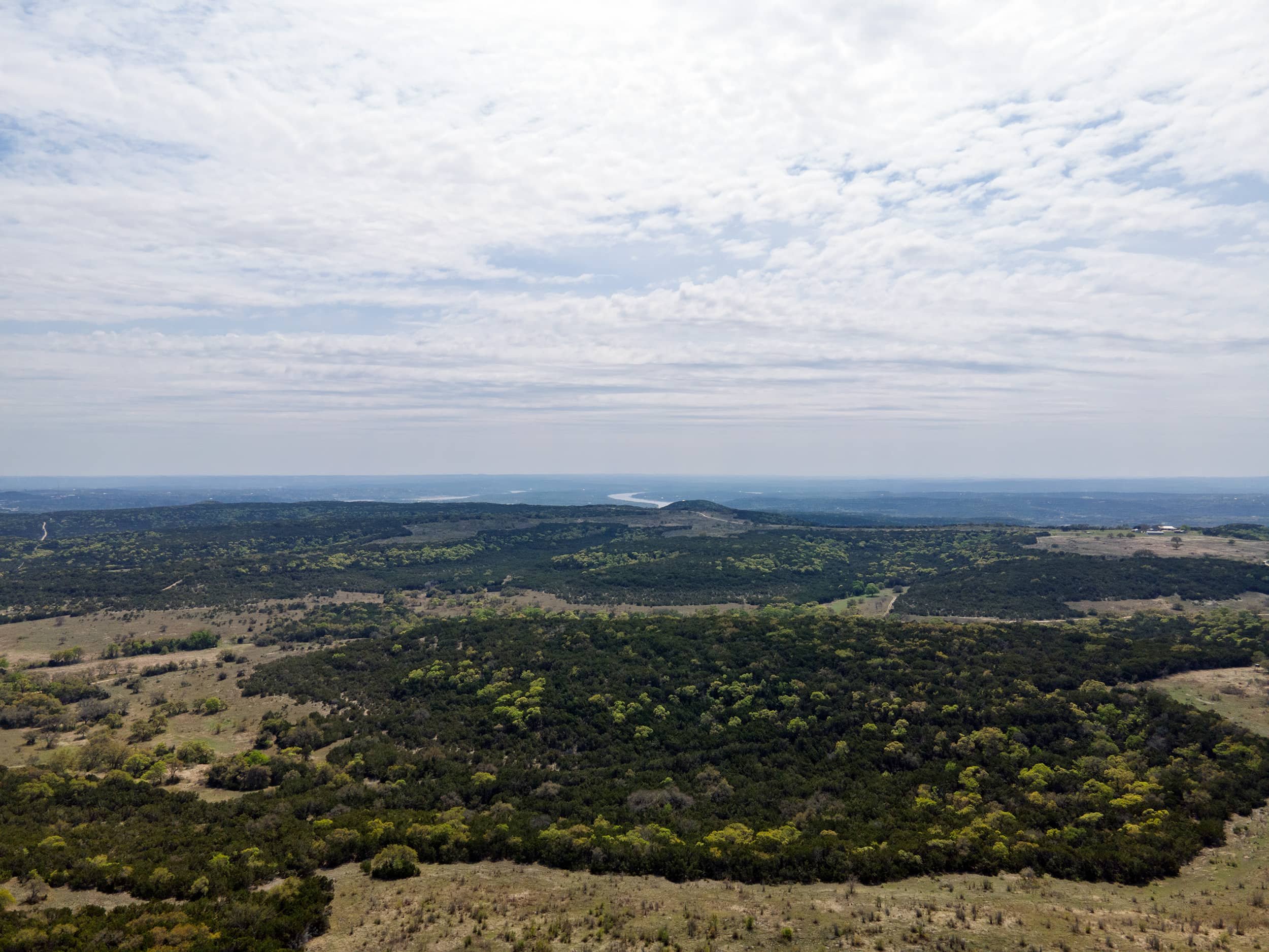 A wide landscape view of a rural area with rolling hills, patches of trees, and a river in the distance under a partly cloudy sky.