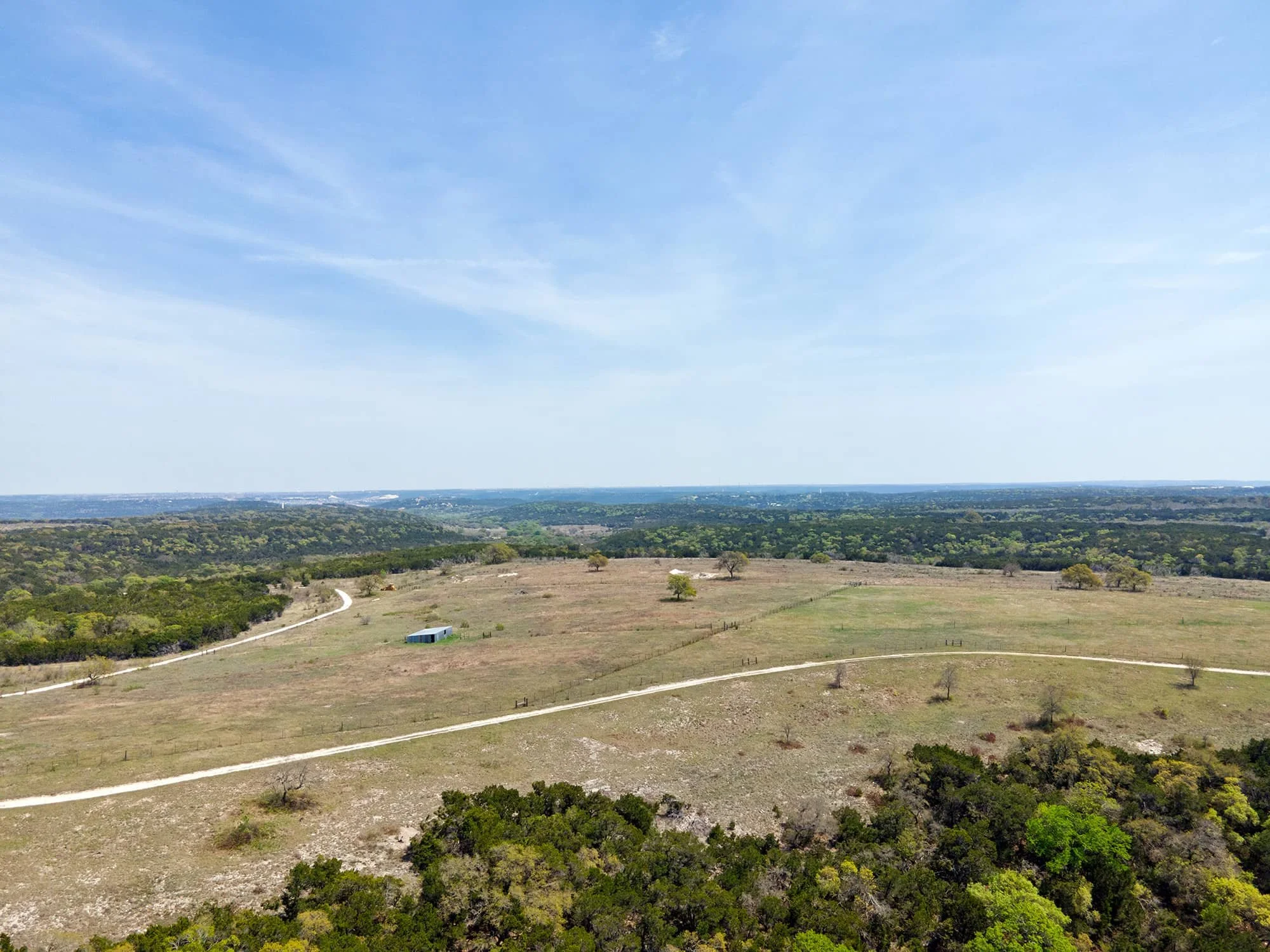 Elevated view of a rural landscape with green fields, scattered trees, a winding dirt road, and a small blue building, under a partly cloudy sky.