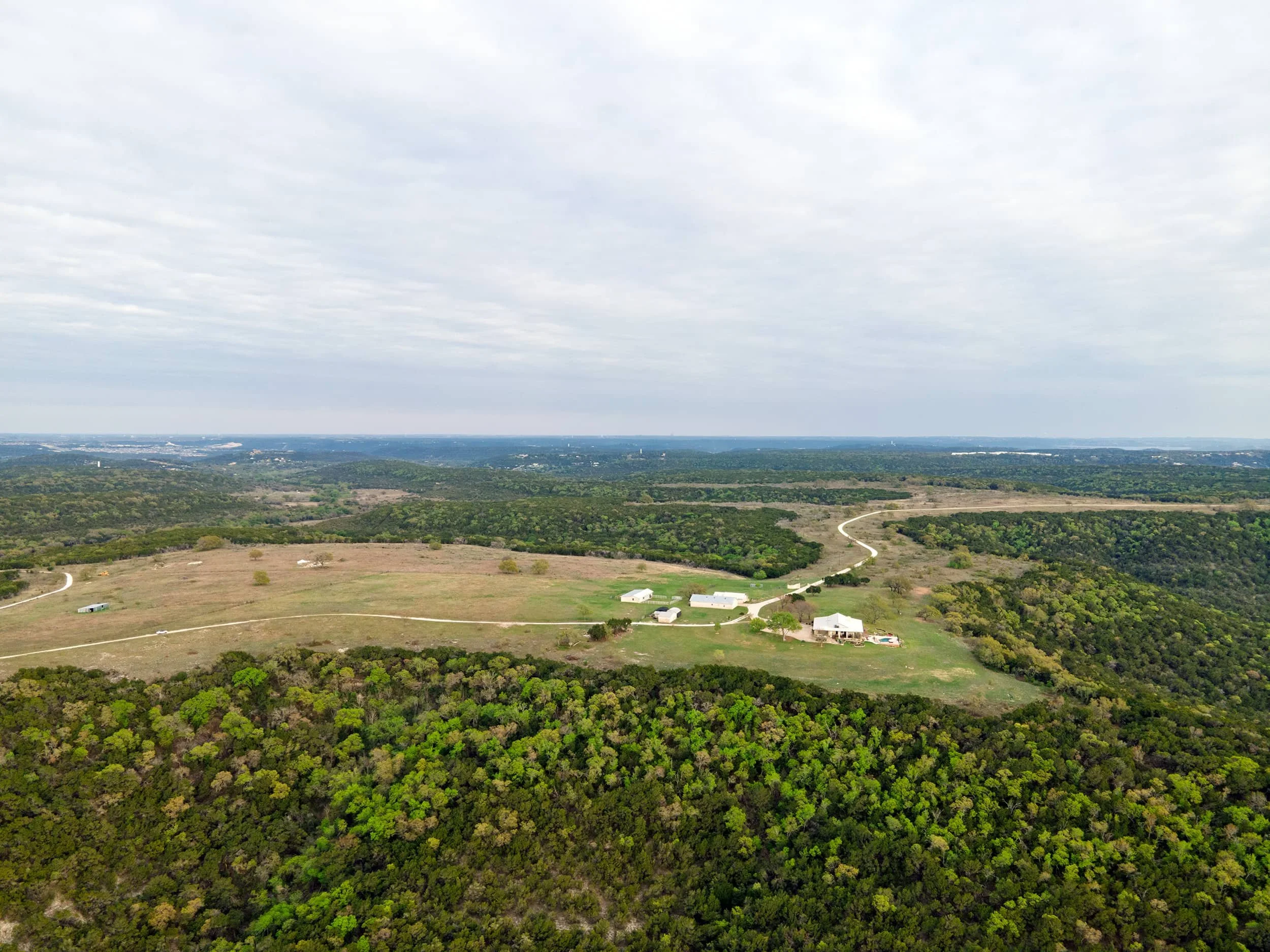 A sprawling landscape with green trees in the foreground, open fields and farm structures in the middle, and a distant city skyline under a cloudy sky.